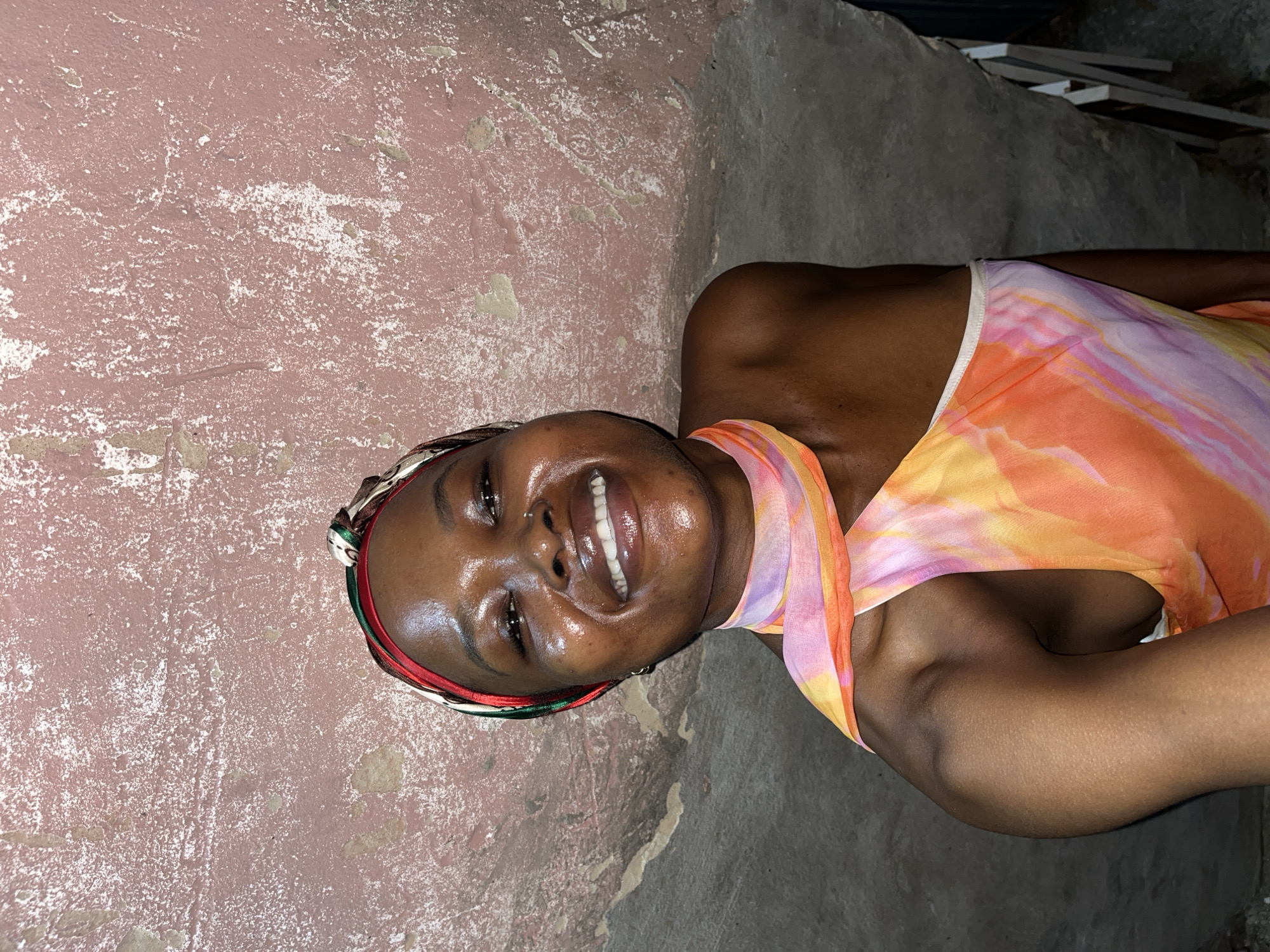 A smiling dark-skinned woman with a headscarf and a colorful orange, yellow, and pink dress is lying on the ground. The background consists of a weathered pink wall and a gray concrete floor or surface. Some debris or building materials are visible in the upper right corner. The location appears to be an urban environment, potentially Ado, Nigeria.
