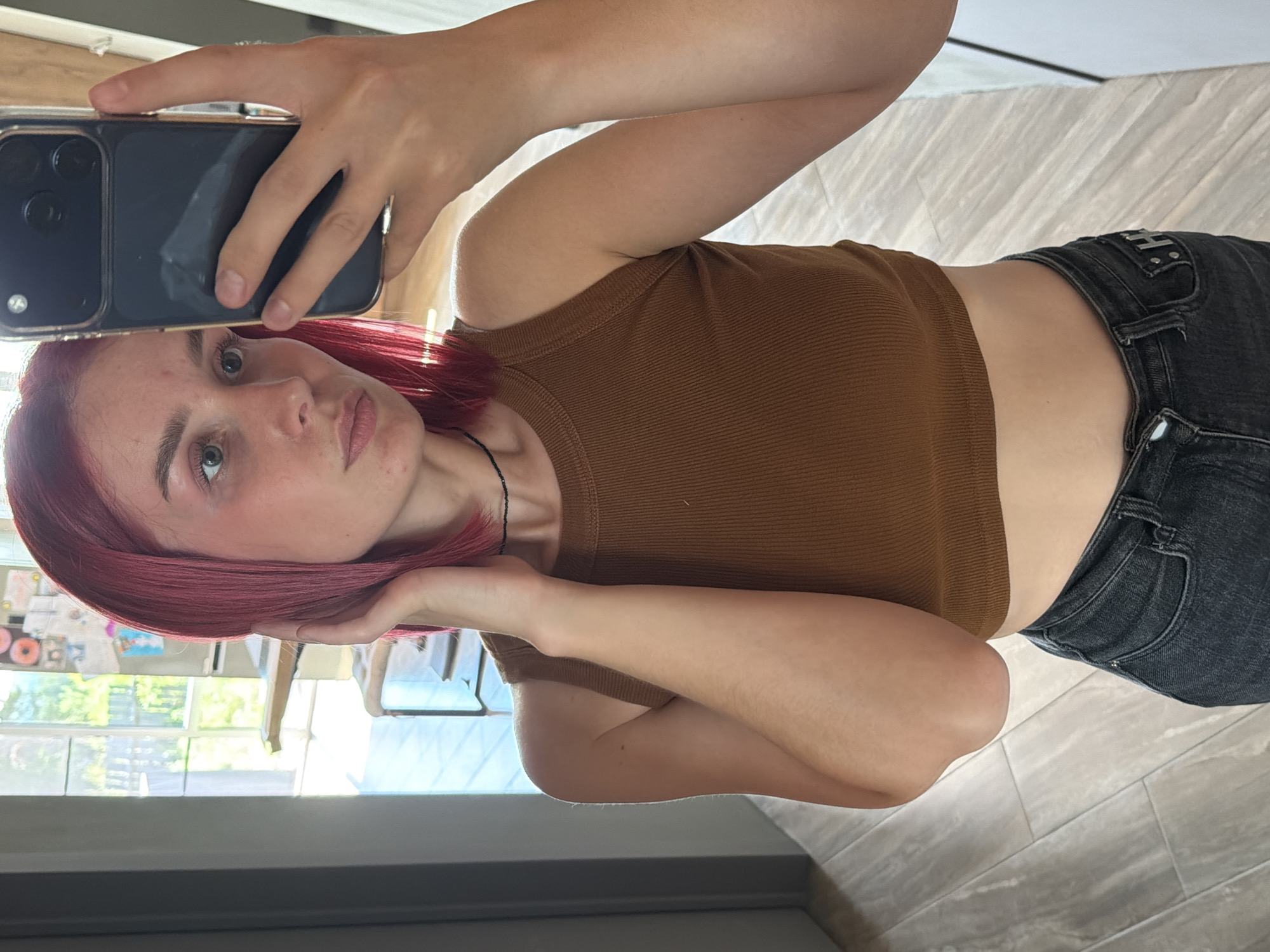 The scene, understood to be located in the city of Chaikovsky, Russia, depicts a young adult female taking a mirror selfie indoors. The individual has shoulder-length, straight red/magenta hair, parted on the left side of her head. Her skin tone is light, and she has visible green/hazel eyes, light eyebrows, and naturally pigmented lips, with some minor facial blemishes present. She is wearing a sleeveless, ribbed brown crop top and dark grey denim jeans, visible from the waist down. A thin black beaded choker necklace adorns her neck. Her right hand is extended, holding a dark-colored smartphone, likely an iPhone due to the triple camera array, encased in a clear or translucent cover with a metallic gold-colored frame, pointed towards her. Her left hand is raised to the side of her head, gently touching her hair near her ear. The background shows an interior space with a large window admitting significant natural light, through which blurred green foliage and distant outdoor structures are visible. The flooring is composed of light grey/beige planks, possibly wood-effect tiles. Partially visible in the background are light-colored furniture pieces or shelves and blurred wall decor.