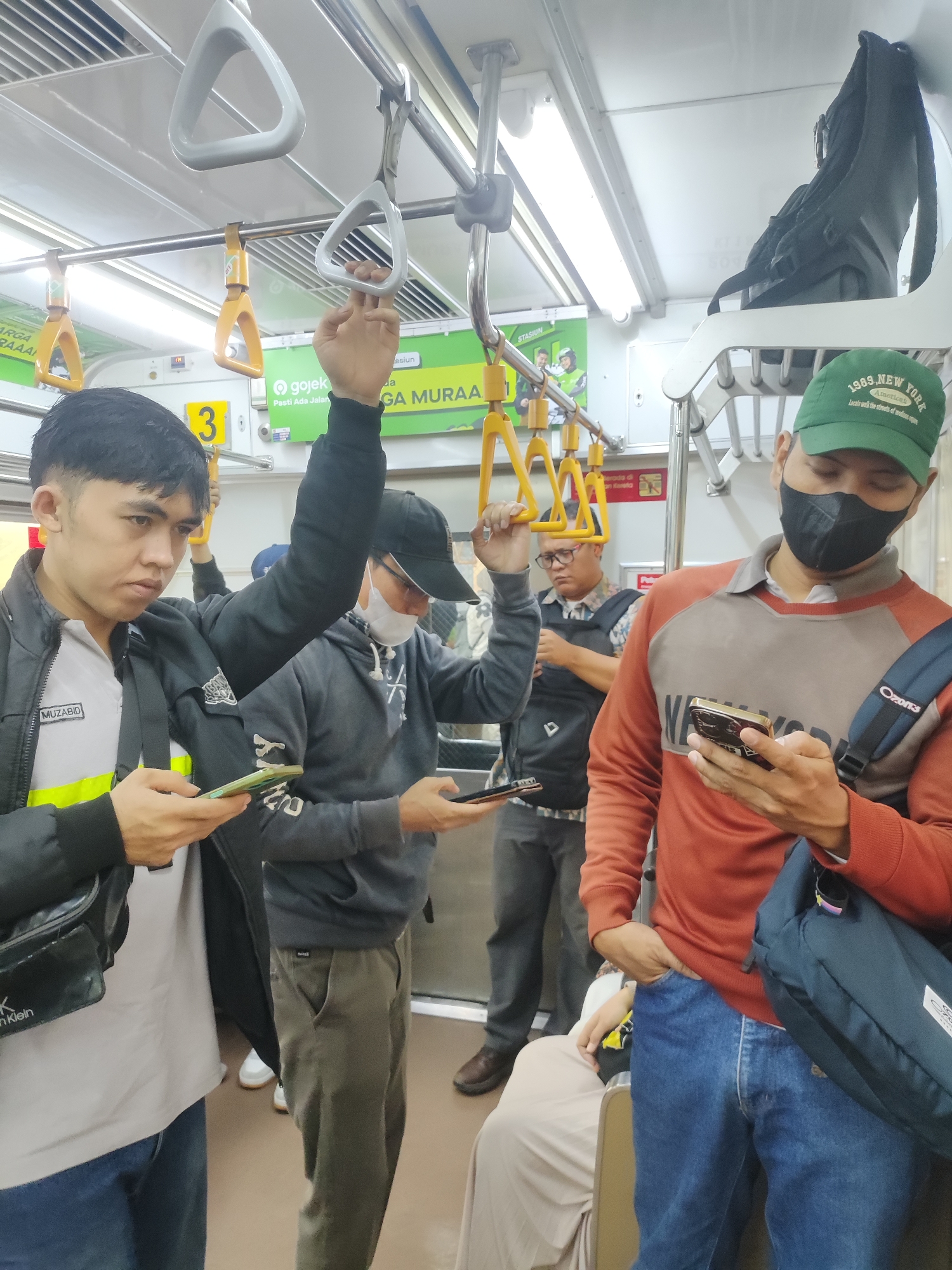 The image captures the interior of a crowded commuter train in Tambun, Indonesia, specifically on the KRL Commuter Line. Several passengers are visible, primarily men, standing and holding onto overhead railings. The lighting is typical of a train carriage, with fluorescent lights illuminating the scene.
In the foreground, a man in a black jacket over a light-colored shirt with a yellow stripe is looking down at his phone. To his right, another man wearing a grey hoodie and a black baseball cap is also holding a phone. Further to the right, a man in a green baseball cap and a black face mask is looking at his phone, dressed in a rust-colored long-sleeved shirt and blue jeans.
Behind them, other passengers are partially visible, some holding onto the yellow and white grab handles that line the ceiling of the train. Advertisements and signage are present on the walls, including a "Gojek" logo and text in Indonesian. The floor appears to be a light brown color. A black backpack is also visible hanging from an overhead rack. The overall atmosphere suggests a typical commute, with passengers occupied with their mobile devices.