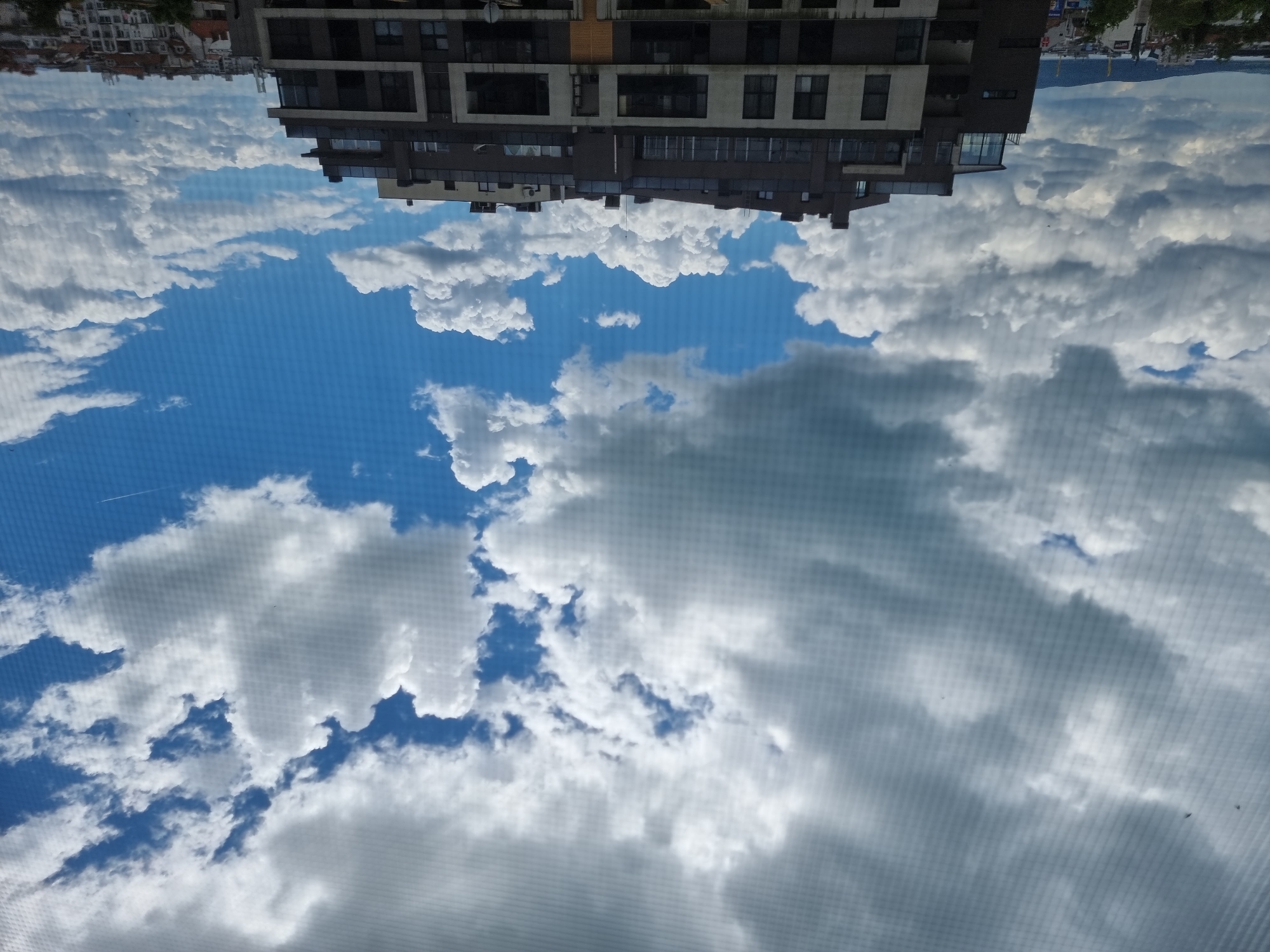 The image depicts an upside-down view of a building in Burgas, Bulgaria, reflected in what appears to be a window. The sky is visible with scattered clouds. The building has a modern architectural style with multiple floors and numerous windows. Other buildings are partially visible in the top left corner of the image. The sky shows a mix of blue and grey, suggesting varying weather conditions.