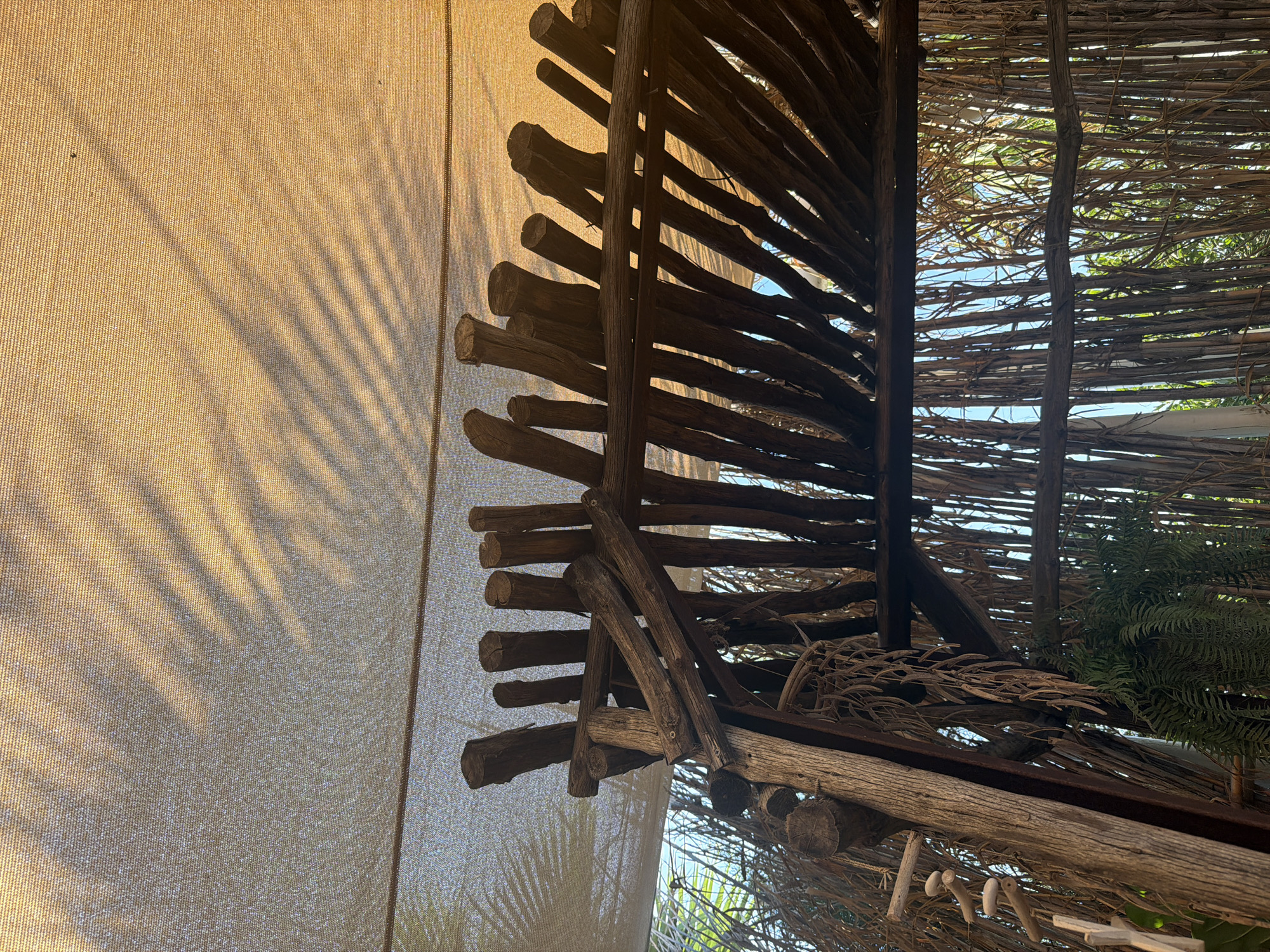 The image captures an outdoor shaded area constructed with natural materials, likely in Protaras, Cyprus, given the context. A light-colored, textured fabric canopy, possibly a shade sail, stretches across the top of the frame, filtering sunlight and casting dappled shadows of foliage onto its surface. Beneath the canopy, a rustic roof structure is visible, built from rough-hewn wooden logs and branches. These wooden elements are arranged in a slatted pattern, creating openings that reveal a view of the surroundings.
The background consists of a dense arrangement of thin, upright wooden poles or bamboo stalks, forming a wall-like barrier. Through these poles, glimpses of lush green foliage and the bright sky can be seen. A fern plant hangs down from the wooden structure on the left side of the image. Several decorative elements, possibly made of wood or shell, hang on strings on the left side of the frame, adding to the bohemian, natural aesthetic. The overall impression is one of a charming, open-air seating or relaxation area, possibly part of a restaurant, cafe, or private garden, designed to offer shade and a connection with nature. There are no people visible in the image.