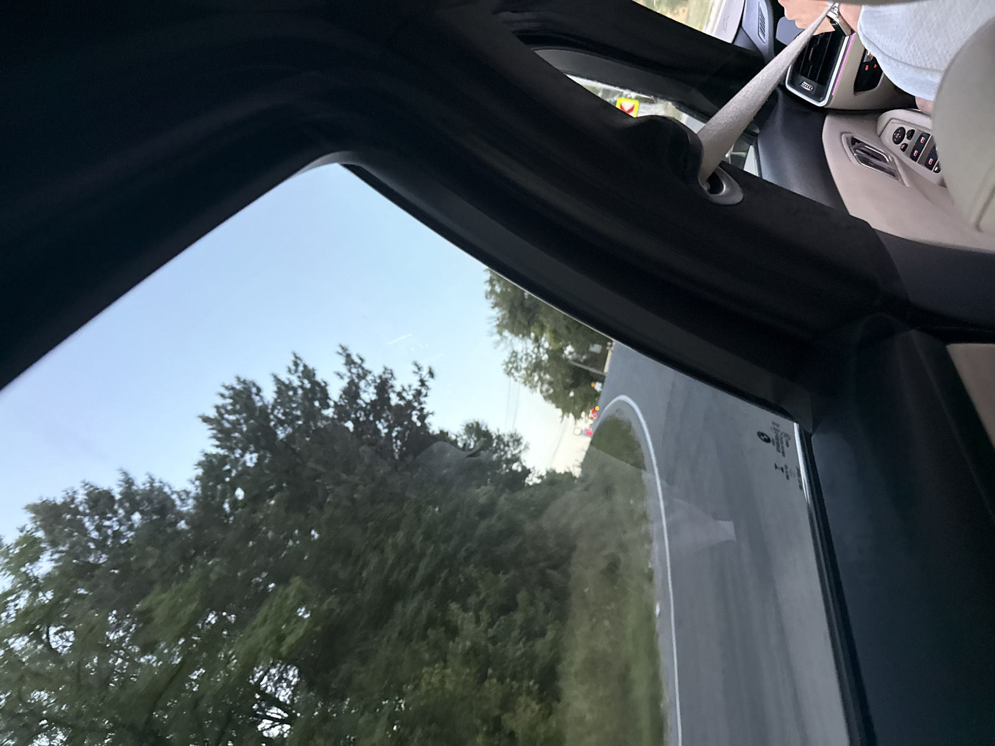 This is a photograph taken from inside a vehicle, looking out the window towards a winding road through a forested area. The sky is clear and light blue, suggesting daytime. The road curves to the right, and on the left side of the road, there is a dense green forest with tall trees. On the right side of the road, further in the distance, there are more trees and what appear to be streetlights or traffic signals.
Within the car, visible through the window, are parts of the interior including a black window frame and trim, and the edge of a tan-colored seat. A person's arm, wearing a watch and holding onto a steering wheel or dashboard element, is visible on the right side of the frame. A seatbelt is also visible, extended across the interior. The photograph has a slightly blurred quality, consistent with being taken while in motion. The image captures a serene, natural landscape with hints of man-made infrastructure (the road and possibly streetlights).