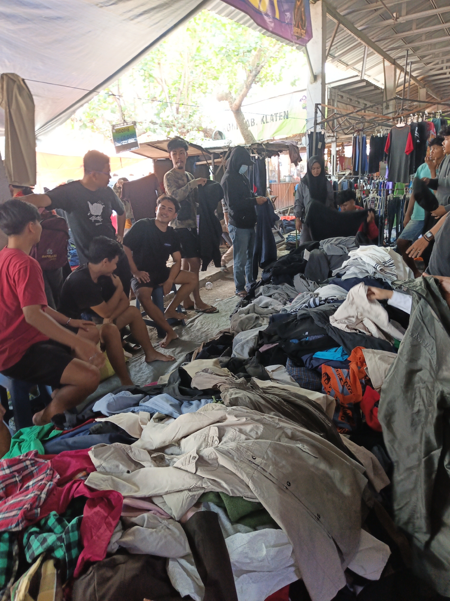 The image depicts an outdoor market scene in Jatinom, Indonesia, likely on a sunny day under a makeshift canopy. A group of young men are gathered around piles of clothing, some sitting on small stools and others standing. Several individuals are interacting with the clothes, either examining them or holding them up. The clothing appears to be second-hand or from a stall, with a variety of items including shirts, pants, and jackets spread out on the ground and on display racks in the background. The environment is somewhat cluttered, with tarps, wooden structures, and other market elements visible. The overall impression is of a casual marketplace where people are browsing and shopping for apparel.