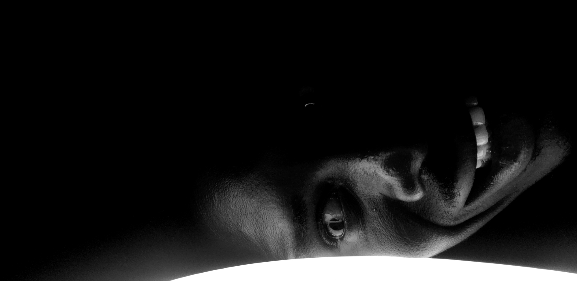 A close-up, black and white image shows a person's face from a low angle. The face is in the lower right of the frame, with an eye visible and the mouth open showing teeth. The rest of the image is black with a white horizontal shape at the bottom. There is no visual information to determine the location in Narayi, Nigeria.