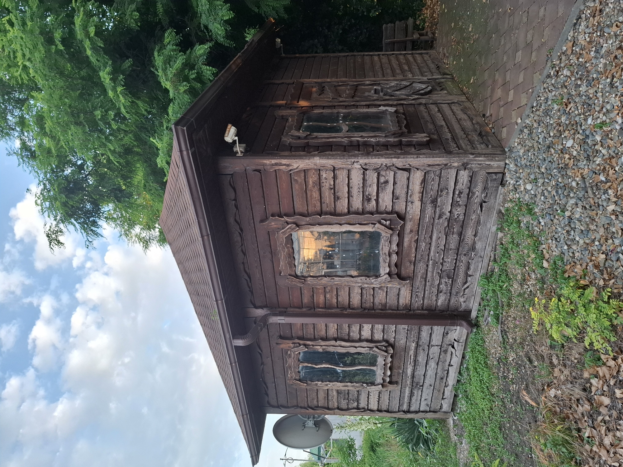 A small, rustic, wooden cabin stands in a yard with a stone walkway and gravel in Korenovsk, Russia. The cabin has a dark brown, shingled roof and dark brown wooden walls. The windows are framed with rough-hewn wood and appear to reflect the sunset. A security camera is mounted on the side of the cabin. To the left of the cabin, a large satellite dish is attached to a pole. Lush green trees are visible behind the cabin. The ground is covered with gravel, fallen leaves, and patches of green grass. A wooden bench is visible to the right of the cabin.