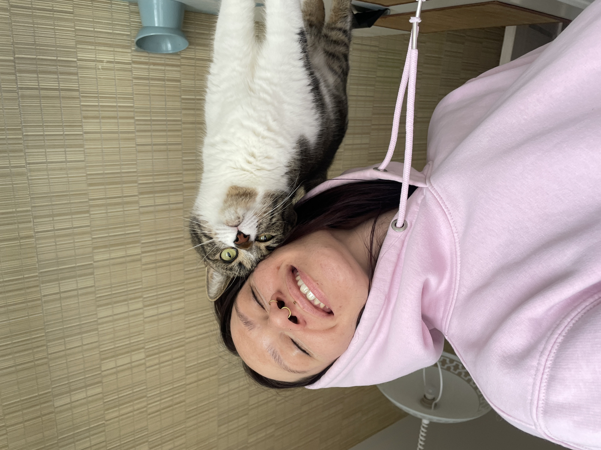 A woman in a pink hoodie is smiling upside down, with a cat positioned above her head, also upside down. The cat is mostly white on its belly and chest, with brown and black tabby markings on its back. The woman has dark hair and a nose piercing. The background includes a textured wall and part of a table. A ceiling lamp is partially visible near the woman's hoodie. The location is likely indoors, but there are no indicators of Izhevsk, Russia.