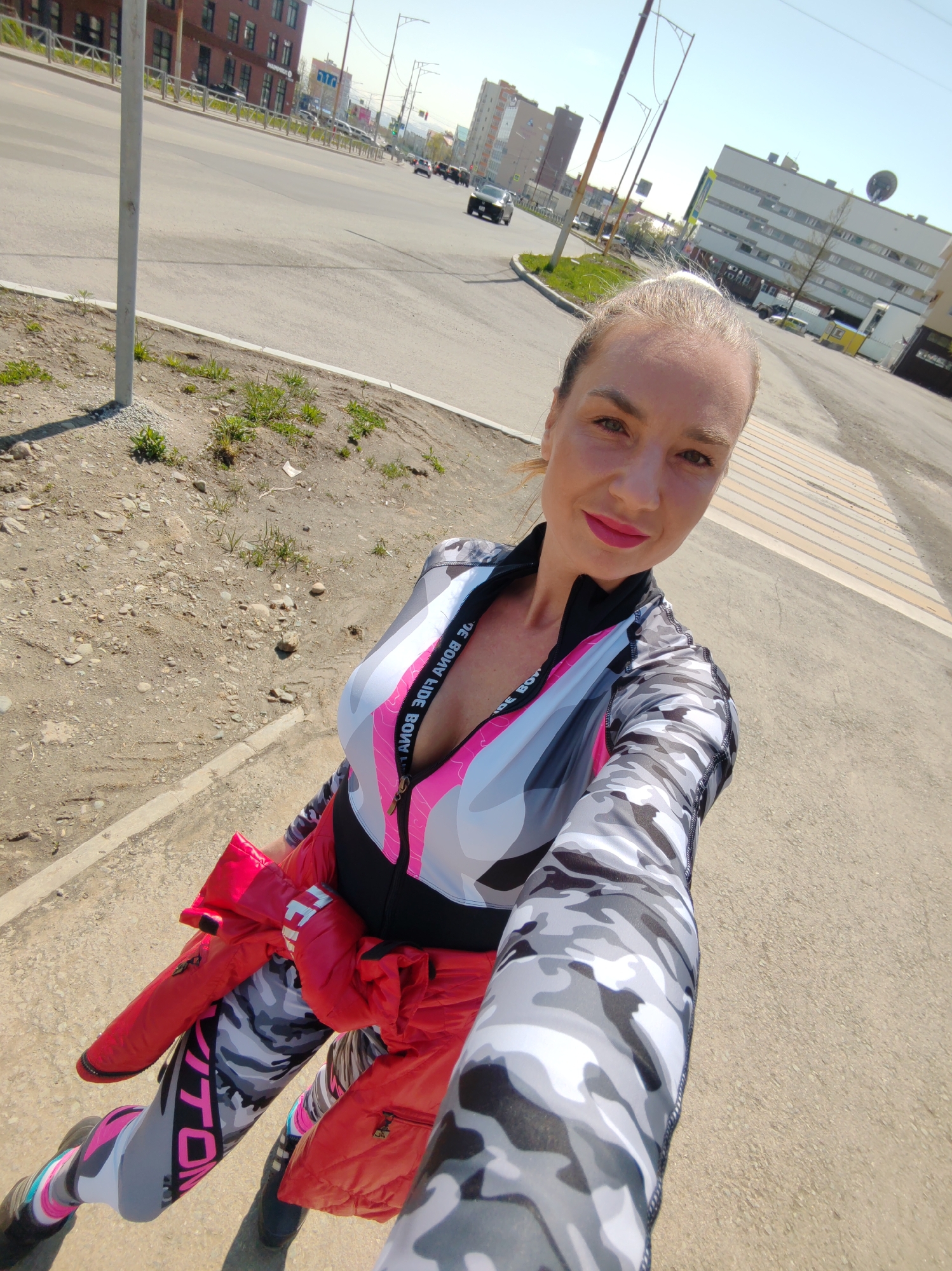 A woman is depicted taking a selfie outdoors on a clear, sunny day in Petropavlovsk-Kamchatsky, Russia. She is positioned on a dirt and gravel patch beside a concrete curb and asphalt road. The woman has light skin, her hair is pulled back, and she is wearing pink lipstick, smiling directly at the camera. Her attire consists of a form-fitting athletic jumpsuit or tracksuit featuring a grey, black, and white camouflage pattern, accented with areas of bright pink and white, particularly on the torso. The garment includes a partial zipper on the front, with the text "BODY FIDE BODY" visible on the trim. A bright red quilted jacket is tied around her waist.
In the background, an urban street scene unfolds under a clear blue sky. To the left, a multi-story red brick building with dark windows is visible. To the right, across the street, a prominent white multi-story building with numerous windows and a large, dark, dish-like structure on its roof is clearly identifiable as a building on Klyuchevskaya Ulitsa (Klyuchevskaya Street), likely associated with the Kamchatka Research Institute of Fisheries and Oceanography (KamchatNIRO) or a related scientific institution, specifically at Klyuchevskaya Ulitsa, 11. Cars are visible driving on the asphalt road, and a pedestrian crossing with white and yellow stripes is present on the pavement to the woman's right. Utility poles and overhead wires traverse the scene. In the immediate foreground to the left, a vertical white pole rises from the ground, surrounded by sparse green vegetation and exposed soil.