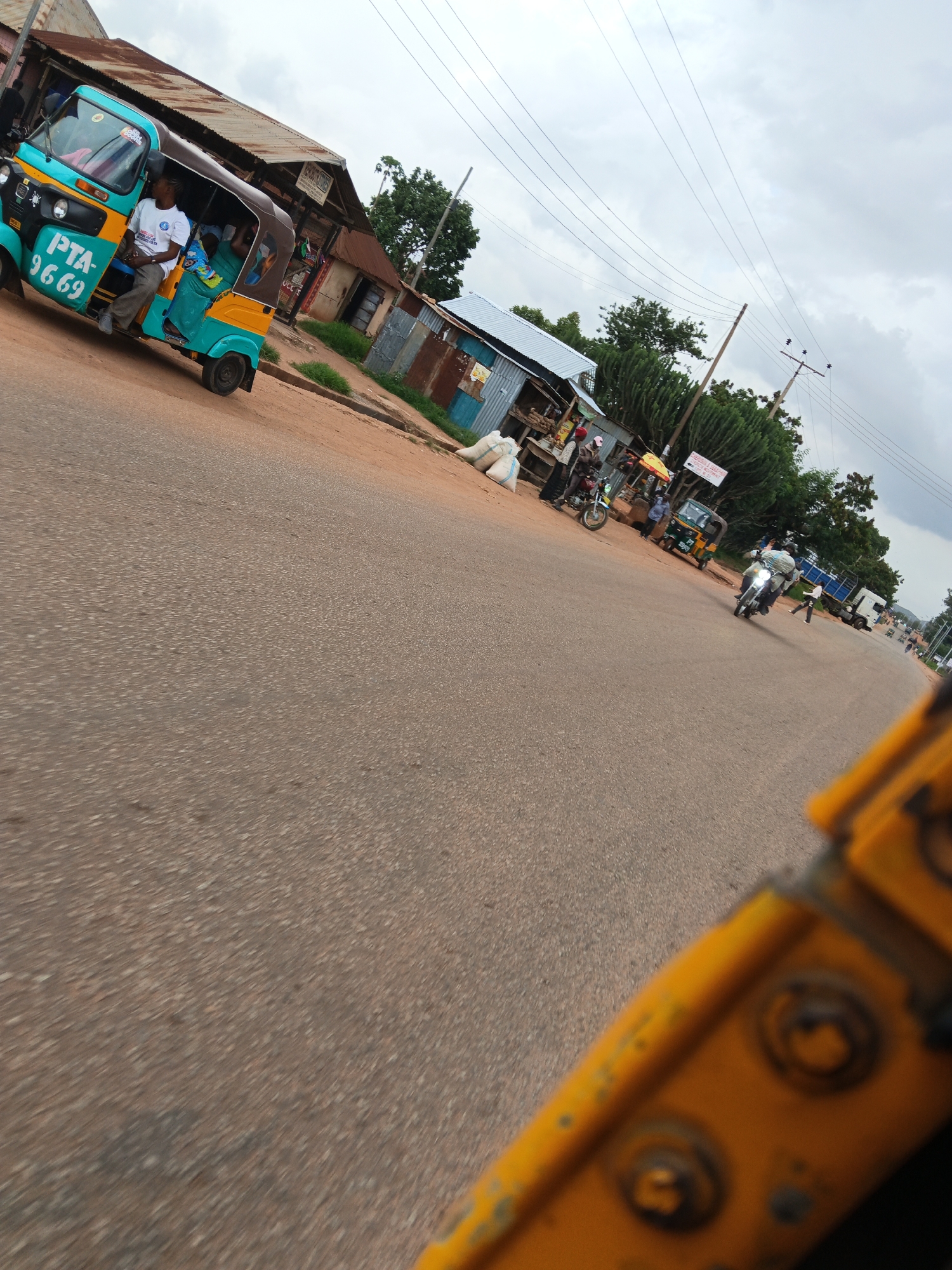 This is a medium-angle, eye-level shot of a street scene, likely in a tropical or subtropical region given the vegetation and architecture. The image is captured from the perspective of a vehicle, possibly a tricycle, as indicated by the yellow element in the foreground. The sky is overcast, suggesting a cloudy or rainy day.
The main subject in the foreground left is a turquoise and yellow tricycle taxi with a brown canopy. A man in a white t-shirt is seated in the driver's position, and another person, possibly a woman in a green outfit, is in the passenger area. The tricycle has "PTA-9669" written in white on its side. To the right of the tricycle, there are several small market stalls with various goods, including large sacks of what appear to be grains or produce. People are standing around these stalls, some on motorcycles.
Further down the road, a man is riding a motorcycle, and another person is walking. A large white truck is parked on the right side of the road. In the background, there are more buildings with corrugated metal roofs and trees, including a prominent cactus-like plant. The road surface is asphalt. The overall impression is of a bustling roadside market in a developing country.