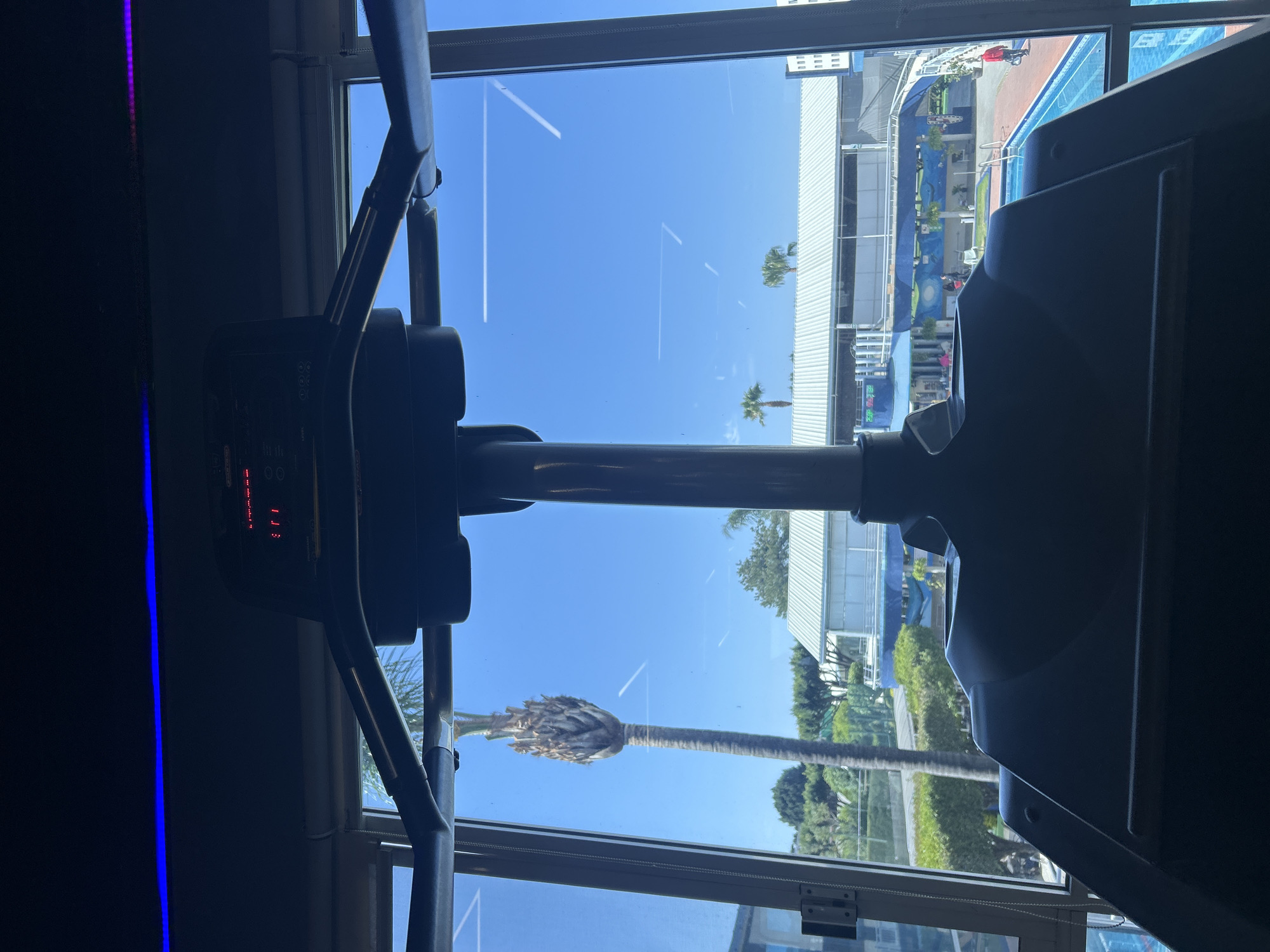 A view from indoors, likely a gym, looking out through a window with a treadmill in the foreground. The window provides a view of an outdoor pool area, a building complex, palm trees, and blue skies. The exact location cannot be determined from the visual information.
