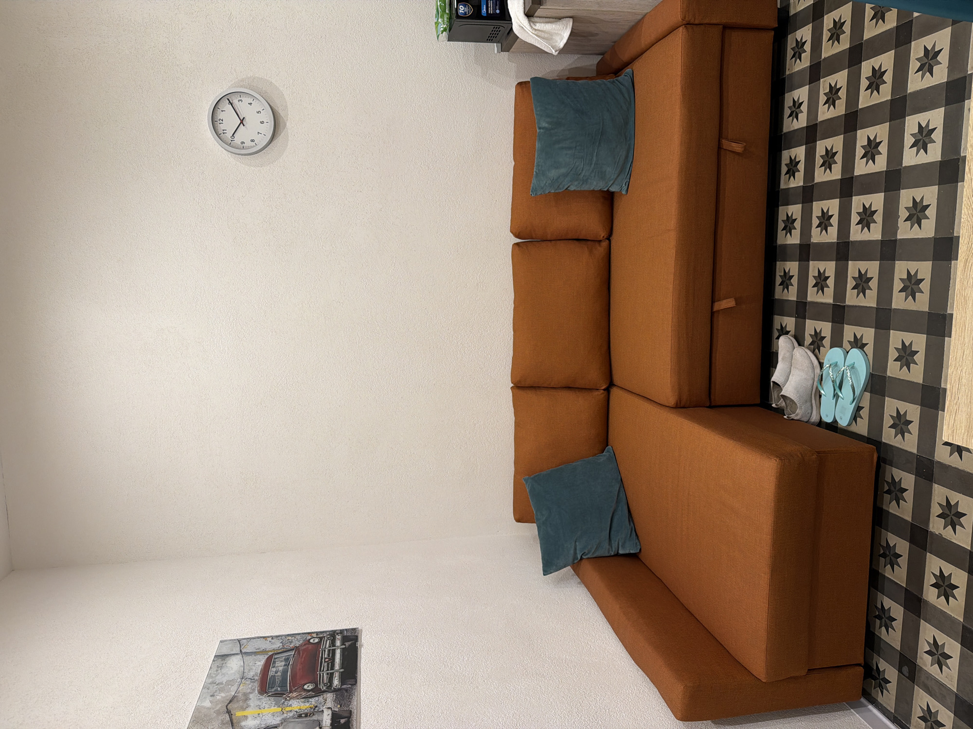 An interior scene depicts a corner of a room with a rust-colored sectional sofa positioned against a textured, off-white wall. Two teal throw pillows rest on the sofa. Above the sofa, a framed picture of a vintage red car is mounted on the wall. To the right of the sofa, a microwave oven is visible on a shelf, with a folded white towel hanging to its left. On the floor in front of the sofa, a pair of light blue flip-flops and a pair of grey slip-on shoes are placed. The floor is tiled with a repeating black, grey, and beige geometric pattern. A round clock with black numbers and hands is mounted on the wall above the sofa. The overall impression is that of a living space or a small apartment. This scene is in the city of Catania, Italy.