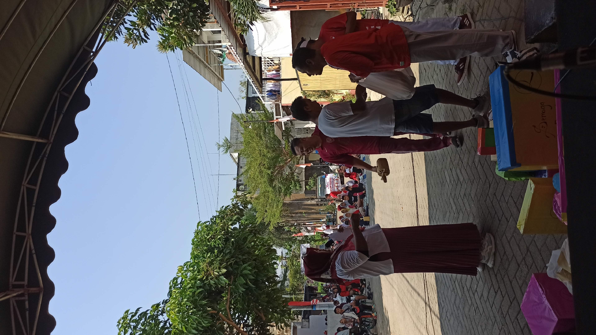 The image depicts an outdoor gathering in Galak, Indonesia, possibly a community event or celebration. In the foreground, several people are interacting. A woman wearing a hijab and a red and white outfit stands on the left, holding papers and facing a group of men. Two men in red t-shirts are on the right, one of whom is handing a white bag to another man. A third man in a burgundy shirt is in the center, holding a wooden object. In the background, a crowd of people, many of whom are seated on the ground, are visible. There are also buildings, trees, and electrical wires, suggesting an urban or semi-urban environment. Some colorful boxes are placed in the lower portion of the image, and a microphone is visible on the right, hinting at a possible performance or presentation. The overall atmosphere appears to be sunny and casual.