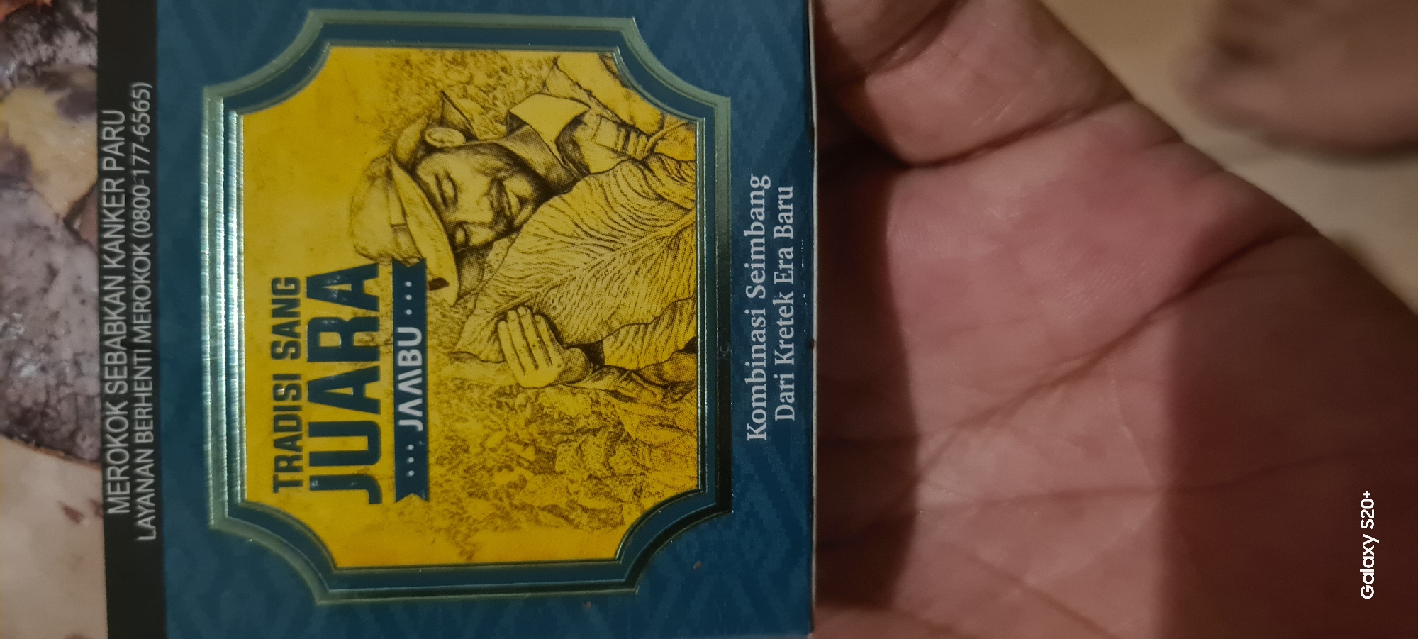 The image shows the front of a cigarette pack, featuring a yellow graphic with a man in a hat holding a large leaf. The text on the pack includes "TRADISI SANG JUARA JAMBU" and "Kombinasi Seimbang Dari Kretek Era Baru". Above the graphic, a warning states "MEROKOK SEBABKAN KANKER PARU" and a phone number for quitting smoking. The cigarette pack is held by a person's hand, with a portion of another hand visible in the foreground, likely belonging to the same person. The background is out of focus. The bottom left corner displays "Galaxy S20+".