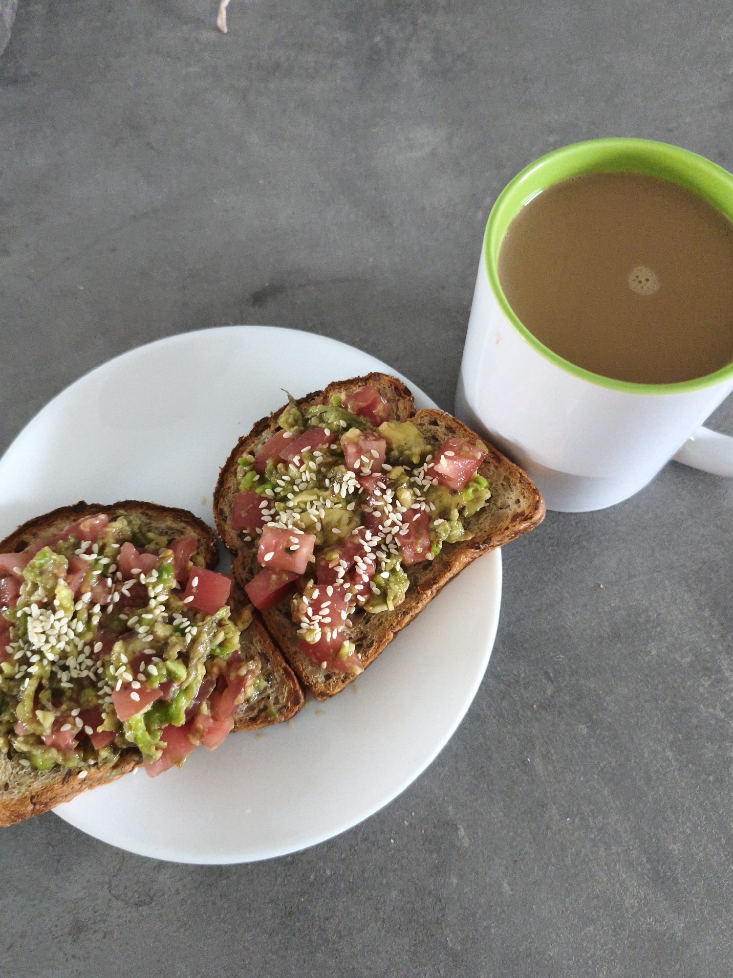 Two slices of avocado toast topped with chopped tomatoes and sesame seeds are arranged on a white plate. To the right of the plate is a white mug with a green rim, filled with coffee. The scene is set against a gray, textured surface, possibly a countertop or table. The location is not identifiable within the city of Brodi, Latvia, as there are no specific landmarks or context clues to suggest this.