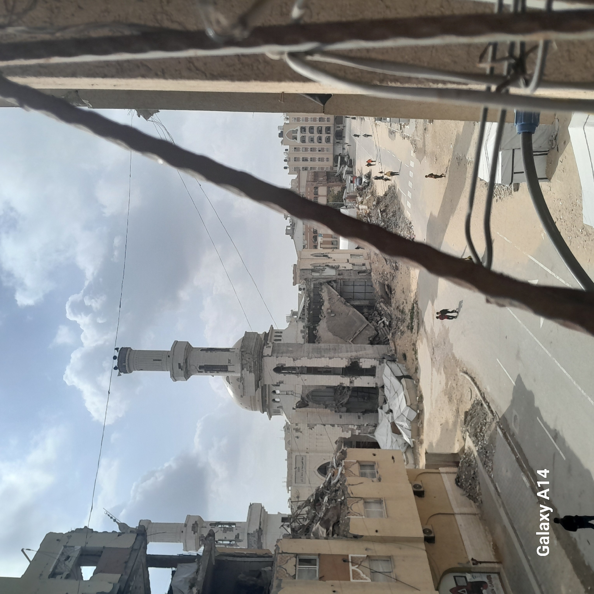 The image shows a partially destroyed city, possibly after a conflict or disaster, as viewed from an elevated position. A mosque with a prominent minaret is visible but damaged, with rubble and debris surrounding it and other buildings. Buildings in the foreground are severely damaged or partially collapsed. A few people are visible on the streets. The sky is cloudy.