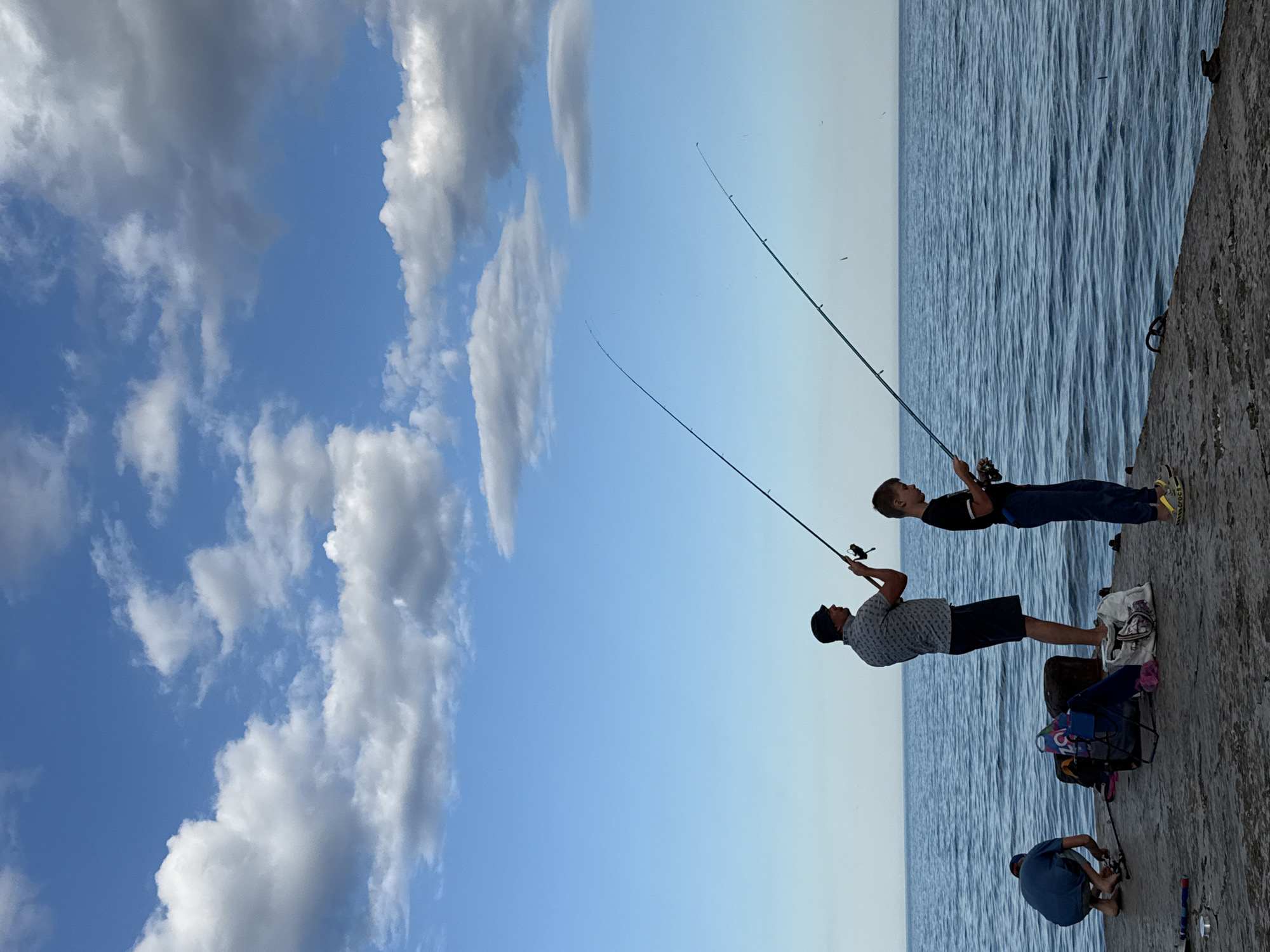 Three individuals are fishing from a concrete pier with the sea and a blue sky with scattered clouds in the background. A man wearing a patterned short-sleeved shirt, dark shorts, and a hat stands holding a fishing rod. Next to him, a boy in a black t-shirt and dark pants also fishes. To the left, a person in a blue t-shirt and shorts is crouched down. Various fishing equipment, bags, and a chair are on the pier near the people. The scene is set on a calm day at a coastal location, likely in Partenit, Ukraine, given the context.