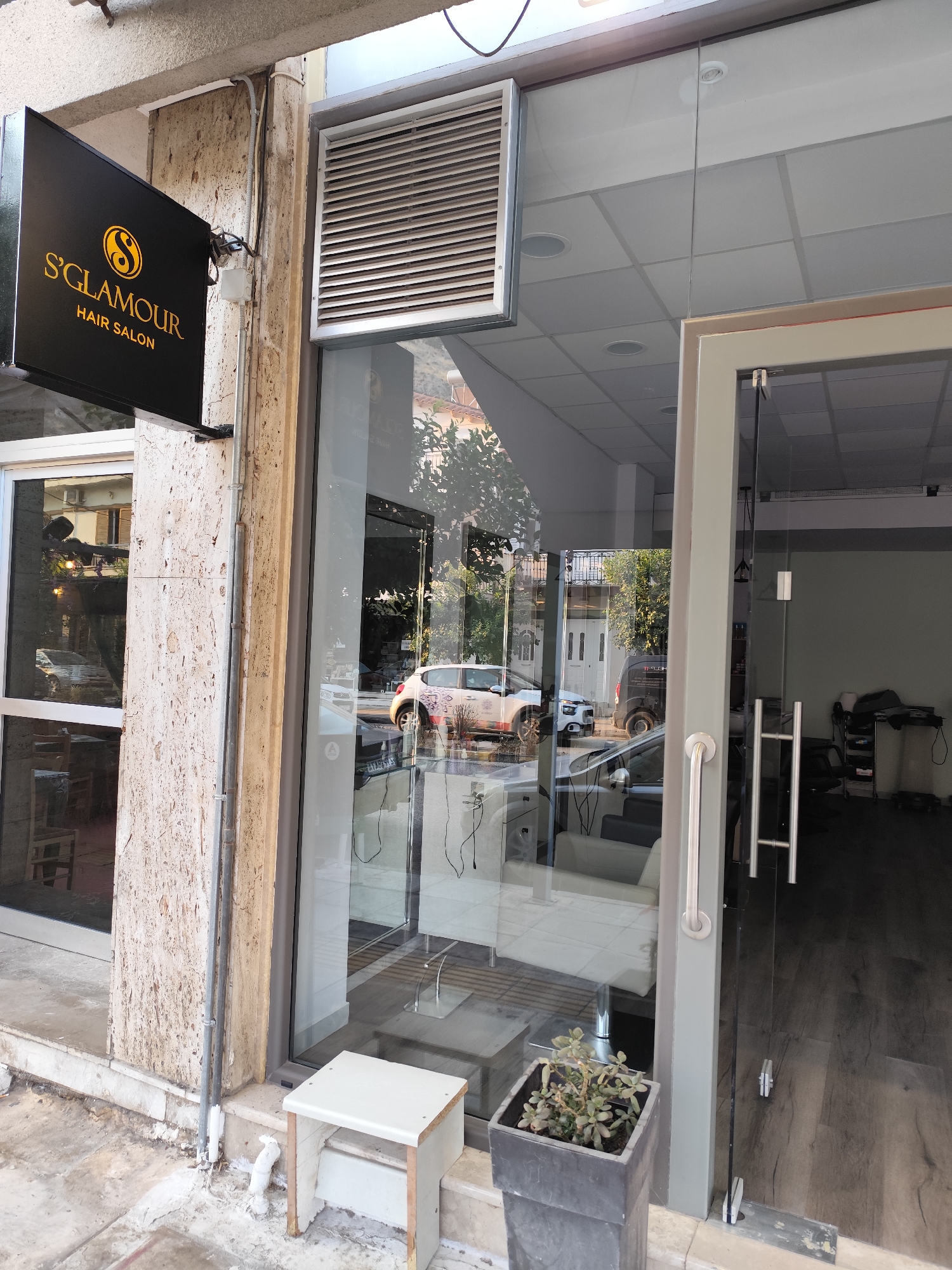 The image shows the exterior of a hair salon named "S'GLAMOUR HAIR SALON" located in the Municipal Unit of Nafplio, Greece. The salon has a glass storefront with a double door entrance. To the left of the entrance, a black sign with "S'GLAMOUR HAIR SALON" written in gold lettering is mounted on the wall. Inside the salon, visible through the glass, are hair salon chairs and equipment. A white stool is placed outside the entrance. The street in front of the salon is visible, with cars parked and a black van. The building exterior is made of stone and plaster.