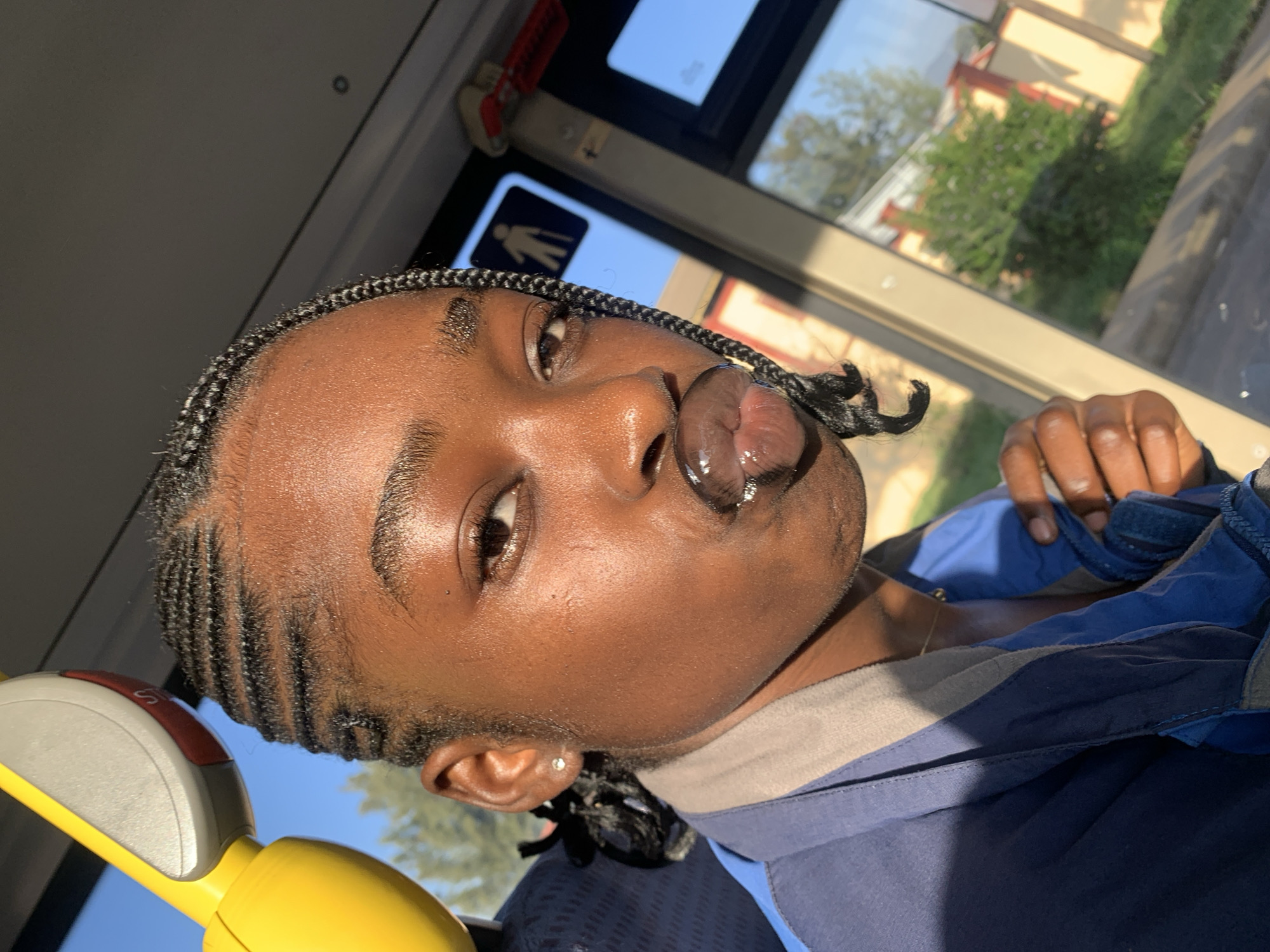 A young Black woman with braided hair is captured in a close-up, selfie-style shot, likely inside a vehicle. Her lips are pursed, appearing to be covered in a glossy substance. She is wearing a blue jacket with grey accents. In the background, sunlight illuminates the interior of the vehicle, highlighting a yellow grab handle and a window that offers a glimpse of a blurred, sunlit exterior with greenery and buildings. The overall lighting suggests it is daytime. The location provided, Rafin Par, Nigeria, is noted.