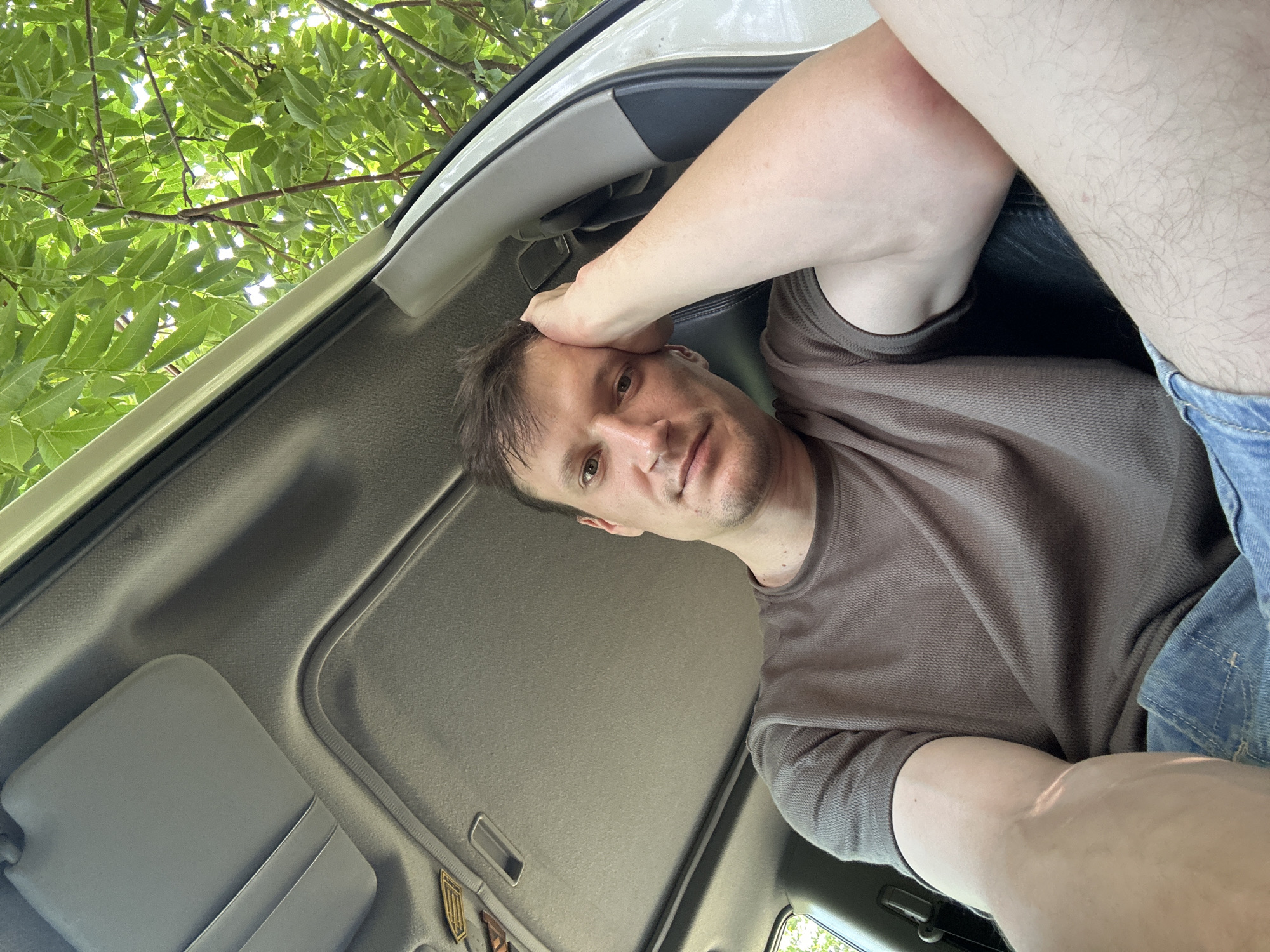 A man with light brown hair and a beard is sitting in the back seat of a car, looking at the camera. He is wearing a brown t-shirt and blue jeans. His right arm is resting on his head, and his left arm is bent at the elbow. The car's sunroof is open, and green leaves from a tree are visible above. The background is blurred, suggesting the car is parked or moving slowly. The overall lighting is natural and appears to be daylight. There is no specific indication of the location being in Makiv, Ukraine.