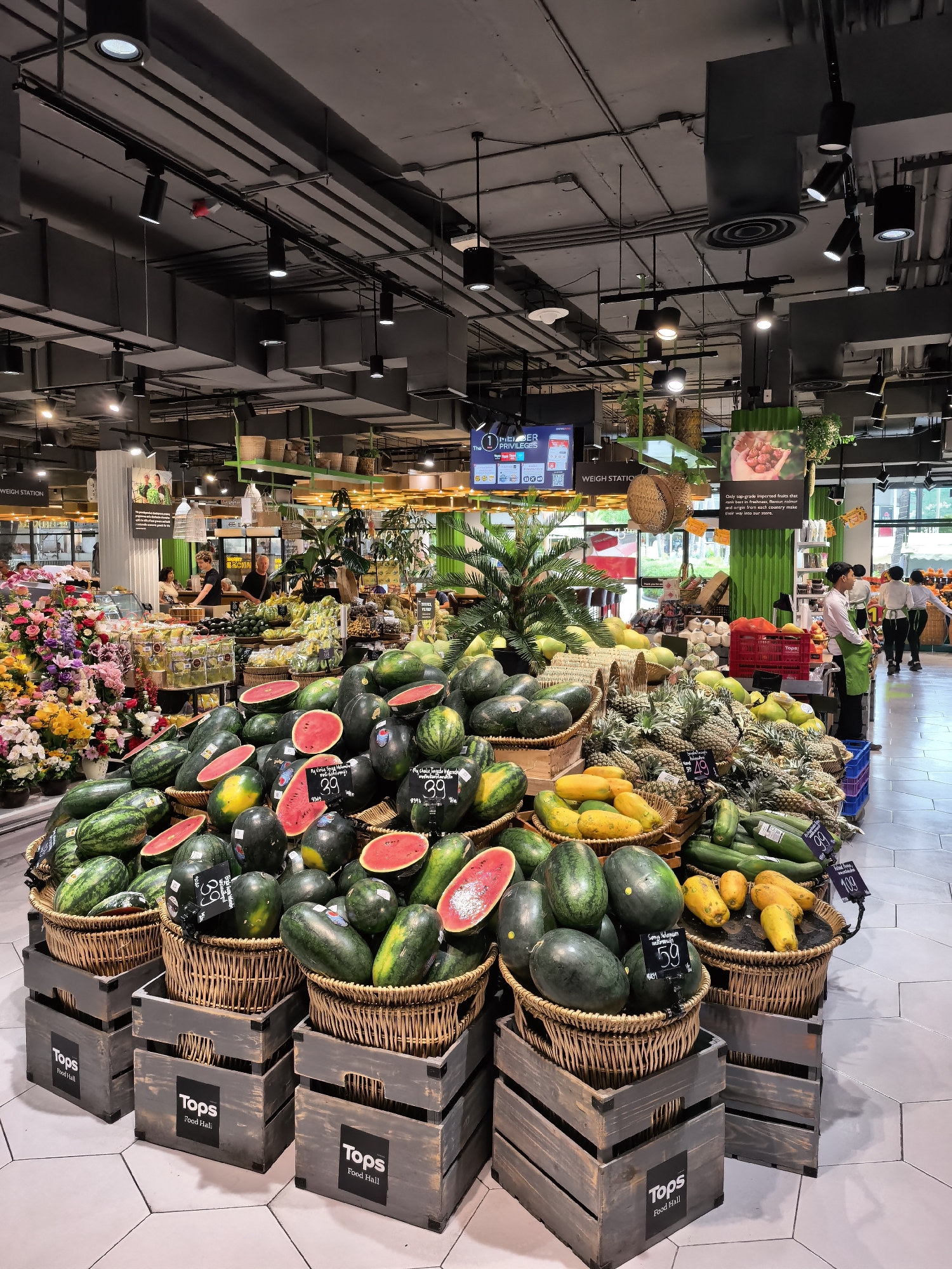 The image depicts the produce section of a "Tops Food Hall" supermarket in the Ko Samui District of Thailand. In the foreground, a large display of watermelons is arranged in woven baskets atop wooden crates. Some watermelons are whole, while others are cut in half, revealing their red flesh. Prices are displayed on tags for the watermelons, showing "39" and "59". To the right of the watermelons, a basket of papayas is visible, with prices of "49" and "99" displayed. Behind the papayas, a large pile of pineapples is displayed. To the left of the watermelons, a colorful display of artificial flowers is arranged.
In the mid-ground, several staff members are visible. A person wearing a green apron is standing near a produce display. Further back, a man and two women are working behind a counter, possibly at a deli or prepared foods section. Other shoppers are also present in the background, browsing the aisles. The supermarket's interior is characterized by its high ceiling with exposed ductwork and a modern lighting system. Large screens displaying promotional information are mounted overhead. The overall environment is clean and well-organized, typical of a modern grocery store.