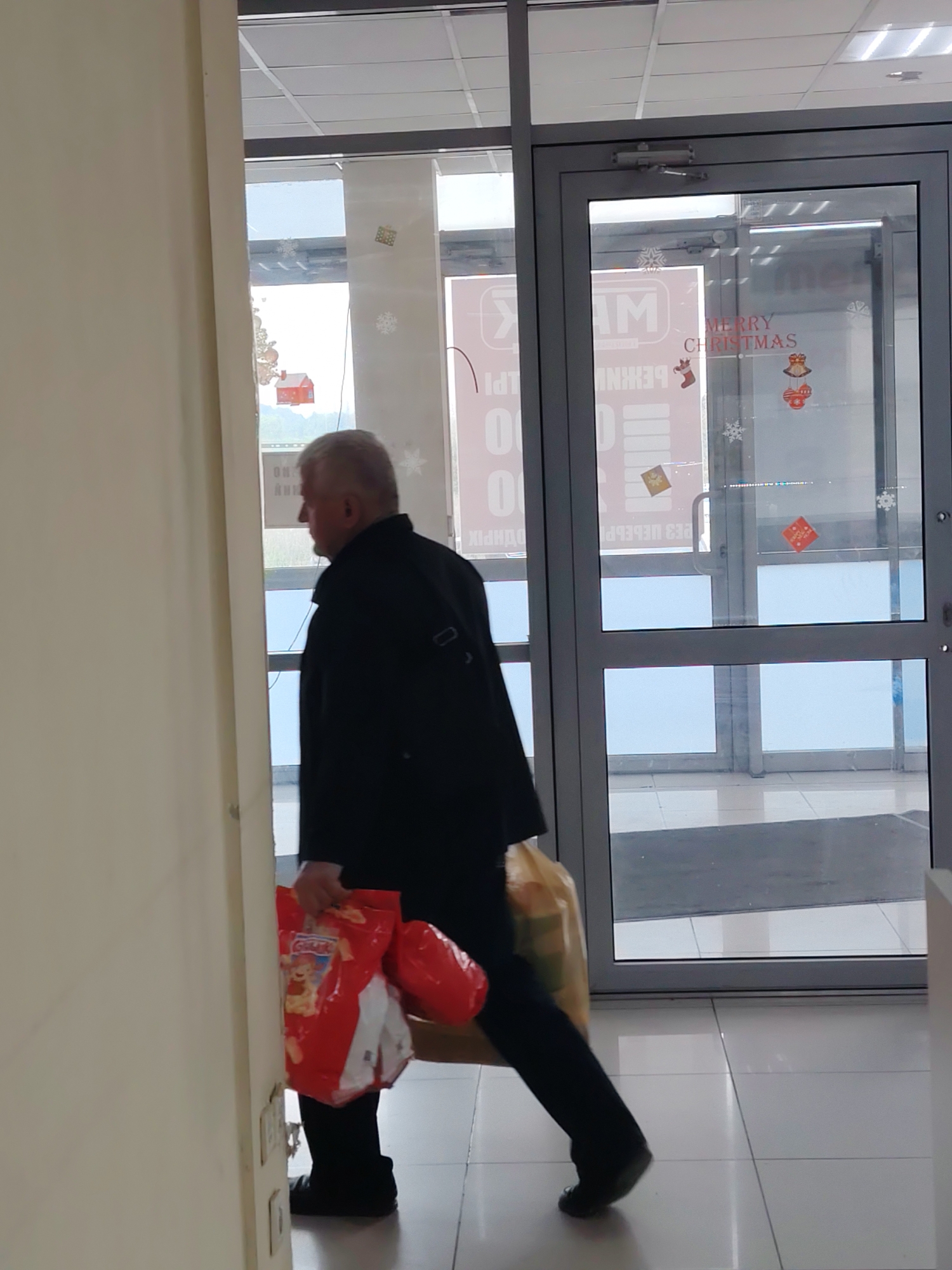 An elderly man wearing a black coat and black pants walks away from the camera carrying two shopping bags. One bag is red and has a cartoon character on it, and the other is a large brown paper bag. He is walking through a doorway with glass doors. The doors have "Merry Christmas" written on them in red letters, along with snowflake and ornament decorations. The floor is tiled in white squares.