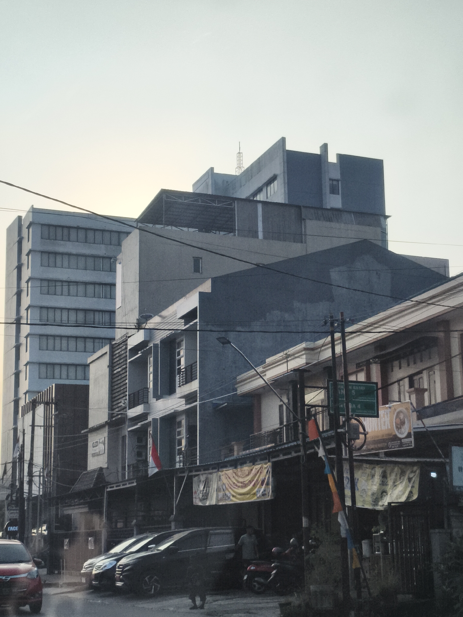 The image depicts a street scene in Samarinda, Indonesia, likely during the daytime or early evening given the lighting. In the foreground, a red car is partially visible on the left, and several other cars are parked or driving on the road. Behind the vehicles, there are multi-story buildings with various architectural styles, indicating a commercial or mixed-use area. Some of the buildings appear to be offices or shops, with signage and awnings visible. A prominent tall, modern building with many windows stands on the left side of the image, contrasting with the more traditional structures in front of it. Overhead, electrical wires crisscross the scene, a common feature in urban Indonesian environments. The sky is overcast, suggesting cloudy weather. A traffic sign with Indonesian text is mounted on a pole, further confirming the location. There are a few people visible, some standing near the buildings and one person walking in the street. The overall impression is of a busy, somewhat cluttered urban streetscape.