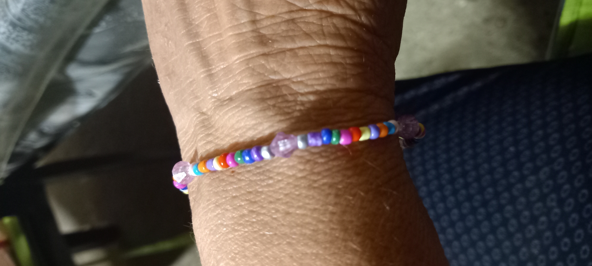 A close-up shot shows a colorful beaded bracelet on a person's wrist. The bracelet is made of small, round beads in various colors including purple, silver, pink, green, blue, orange, and white. There are also two translucent purple charms shaped like flowers or hearts on the bracelet. The wrist appears to be that of an adult, with visible skin texture. The background is out of focus, but appears to be indoors with dark surfaces, possibly furniture or clothing. The image does not provide any information about the location in Iskra, Bulgaria.