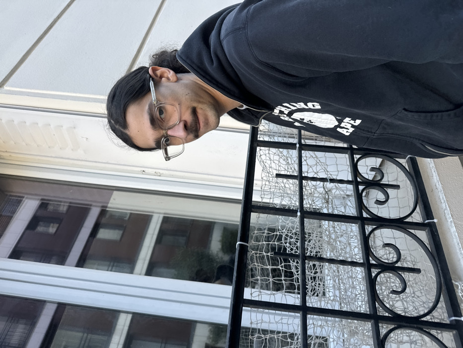 A male subject is depicted in a medium shot, positioned on what appears to be a balcony or ledge in Vincennes, France. The subject has dark, medium-length hair styled back, a thin mustache, and wears clear-framed eyeglasses. He is wearing a dark, zip-up hooded sweatshirt featuring a white graphic logo of a gorilla head with the text "BATHING APE". The subject's head is slightly turned, and he is looking directly at the camera with a neutral expression.
In the foreground, a black wrought-iron balcony railing with decorative elements is present. Attached to this railing is a white, wide-mesh netting, secured with white zip ties, spanning across the visible section of the railing.
The immediate background shows a white building facade with a tall, white-framed window. The window's surface reflects an opposing multi-story building, characterized by multiple rectangular windows and a dark, possibly reddish-brown, facade. A faint reflection of greenery, possibly a tree, is also visible within the window pane.