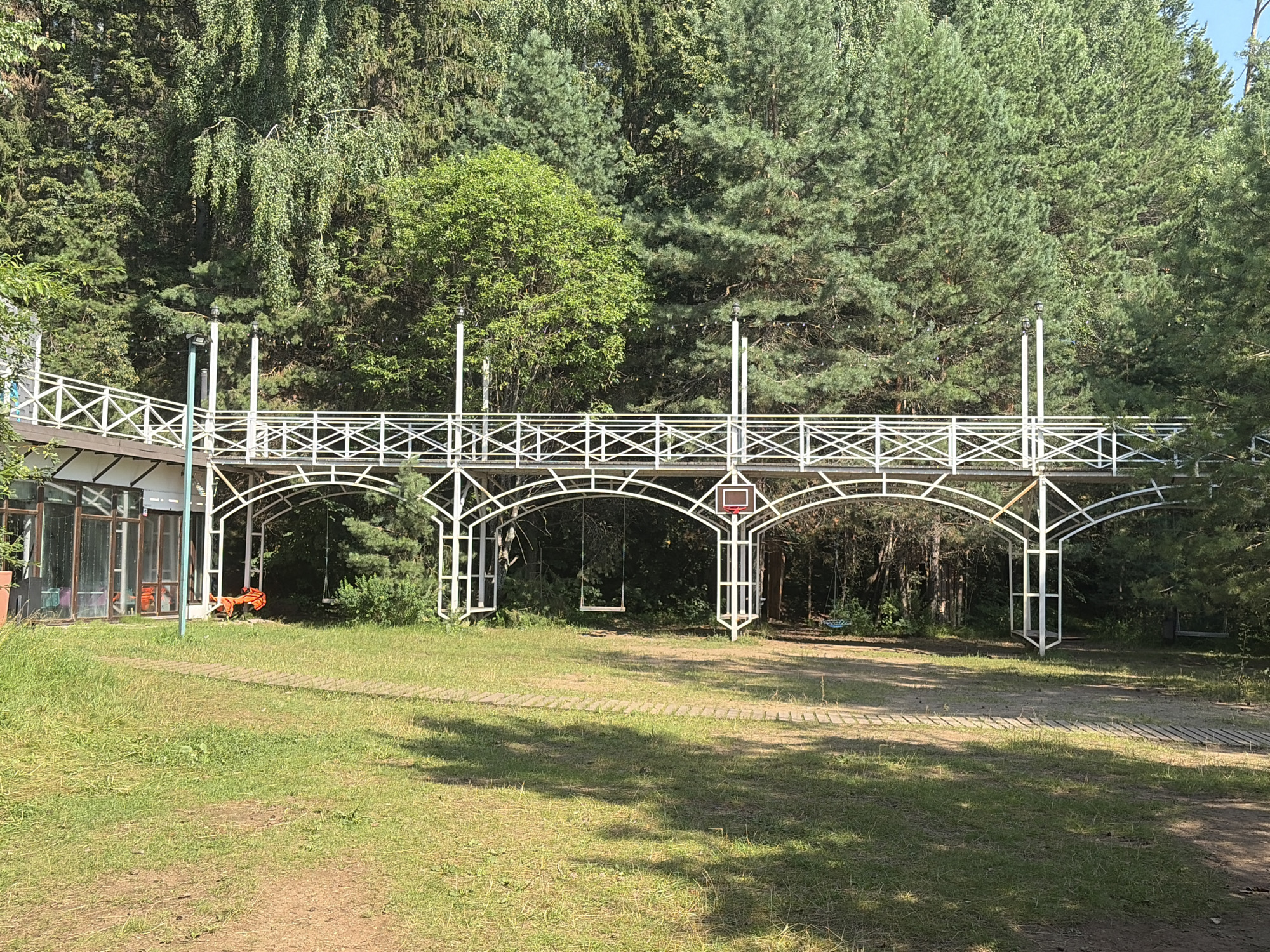 A low-angle, full outdoor shot shows a white, ornate, elevated walkway with arches and a decorative railing, surrounded by lush green trees and grass. The walkway is supported by white, lattice-like pillars and features two hanging swings beneath its structure. On the left side of the image, a glass-enclosed building is visible, with bright orange life vests resting on the ground nearby. The scene appears to be in a park or recreational area, likely in Kostovaty, Russia, given the naturalistic setting and the presence of playground elements. The sky is clear and blue, indicating a sunny day.