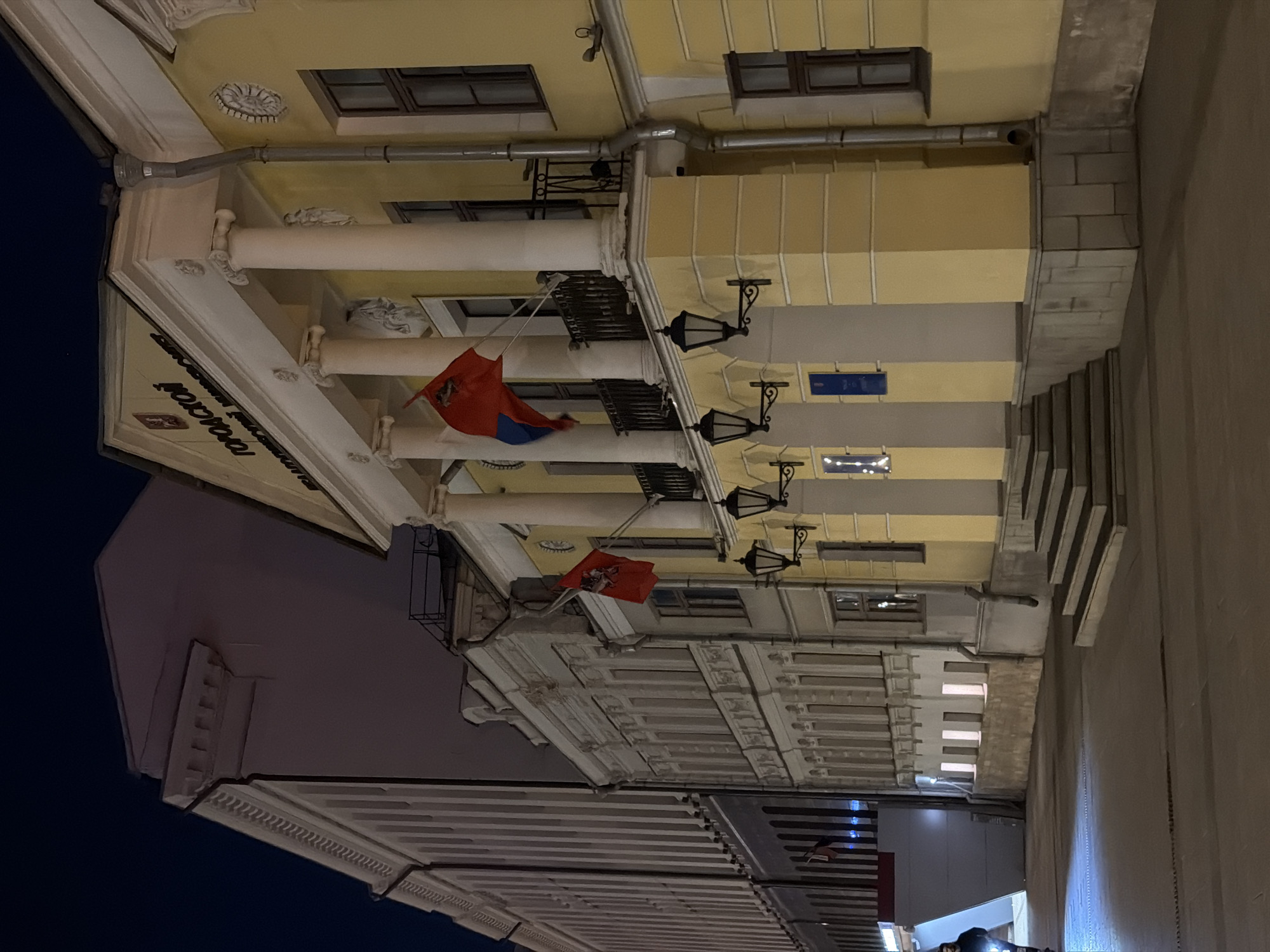The image depicts a street scene in Moscow, Russia. The architecture features classical elements such as columns and decorative moldings on yellow buildings. Russian flags are displayed on the buildings. Street lamps are mounted on the sides of the buildings. The sky is dark, suggesting it is either dusk or night. There are stairs and a wall that appear to lead upwards on the right side of the image.