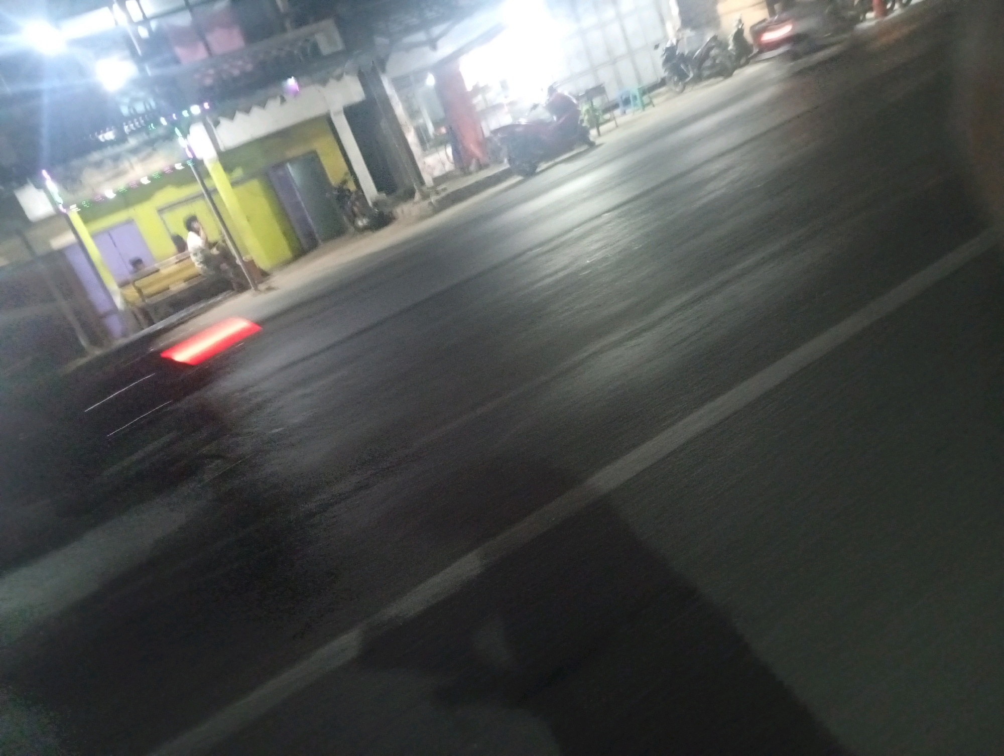 The image depicts a street scene at night, likely in Banowan, Indonesia, given the context. The perspective suggests the photo was taken from a moving vehicle, resulting in motion blur, particularly on the road surface. On the left side of the image, a building with a brightly lit facade is visible, adorned with string lights. A person is seated on a bench in front of this building. Further down the street, other buildings are present, with some appearing to be shops or residences. Several motorcycles are parked or in motion, one of which has a visible red taillight. The road surface is asphalt and appears wet, reflecting the ambient light. The overall atmosphere is that of a bustling street at night.