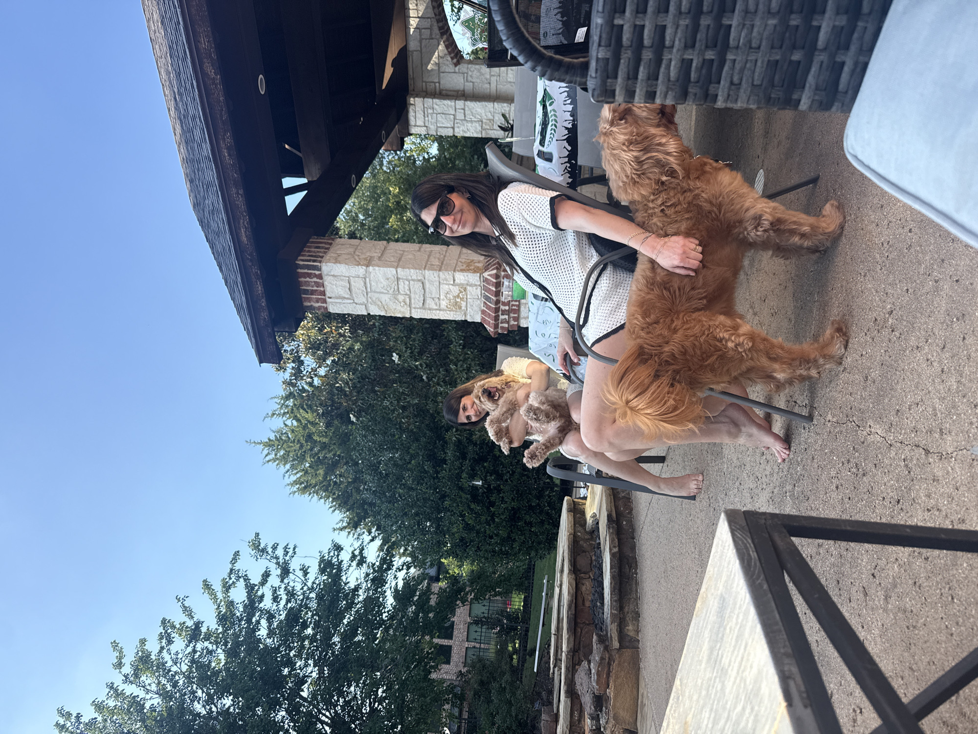 A woman wearing sunglasses and a white dotted sweater sits outside with two golden doodle dogs in Southlake, Texas, USA. One dog is sitting on her lap and another is standing next to her chair. The woman sits under a covered patio. A table is next to the chair. A stone fire pit is in the background. Trees are visible beyond the patio.