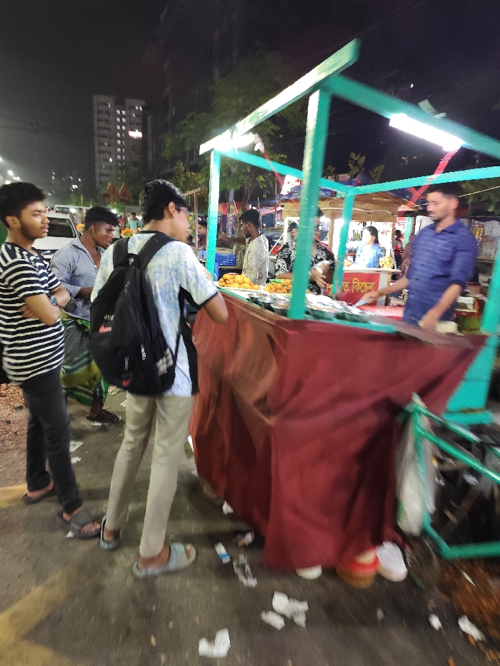 The image shows a street food stall in Dhaka, Bangladesh, located at coordinates 23.78 latitude and 90.374 longitude. The stall is a wooden cart with a red tarp covering the sides. Several people are gathered around the cart, including three men in front of the cart, a woman standing behind the counter, and two men in the background. One man is wearing a black backpack and a blue and white shirt, while another man is wearing a striped shirt. The woman behind the counter is wearing a white shirt and a white head covering. The cart is selling various items, including food and drinks. There are a few pieces of trash on the ground. The environment is dimly lit, suggesting that it is nighttime. The scene is bustling and active, with people interacting with the food stall.