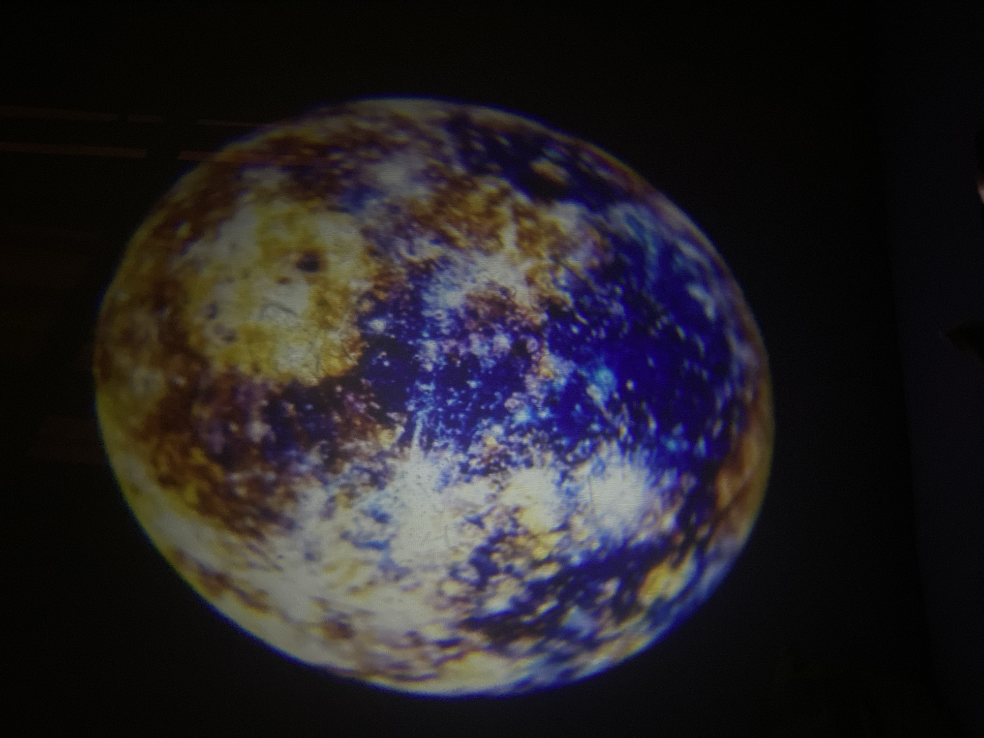 The image shows a projection of a celestial body, likely a planet or moon, on a dark surface. The object has a mottled appearance with various colors including yellow, brown, white, and blue. The dark background appears to be a wall. There are no people visible in the image. The location is unknown, and there is no evidence to suggest it is in Saint Petersburg, Russia.