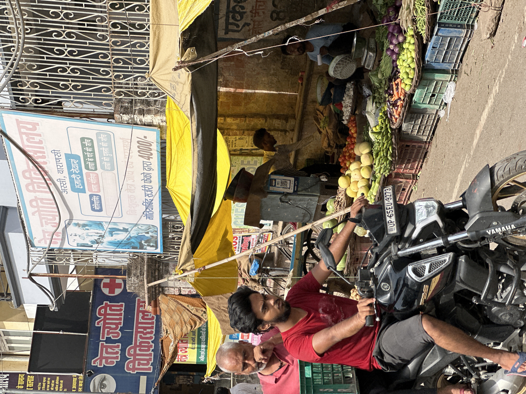 The image shows a street scene in Varanasi, India, with a man on a motorcycle in the foreground. A fruit and vegetable stand is situated under a yellow awning, with vendors and produce visible. Street signs in Hindi advertising eye care services, including "Netra Jyoti" (eye light) and "Jyoti Nidanayala" (Jyoti Eye Clinic) are also visible. The motorcycle's license plate reads "UP-65EU-9273". Two men are on the motorbike, one older and one younger, with the younger man driving.