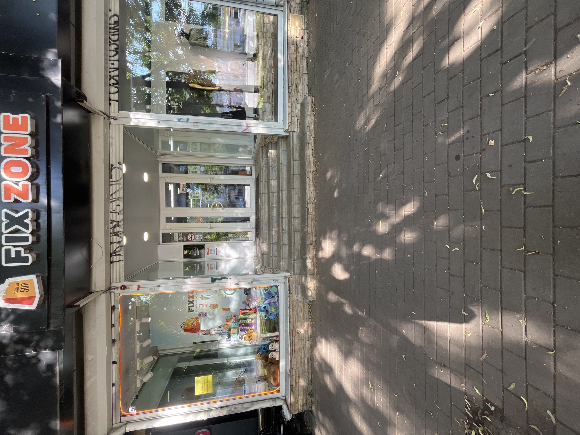 The image shows the storefront of "FIX ZONE", a store located in Voronezh, Russia. The storefront features a large, illuminated sign that reads "FIX ZONE" in orange lettering. The store has large glass windows displaying merchandise. To the left, the window showcases stuffed animals and various products. To the right, the window displays clothing and mannequins. The entrance to the store is in the center, with a set of stairs leading up to it. The building is situated on a brick-paved sidewalk, with shadows from trees cast across it. The overall scene is that of a commercial street in a city.