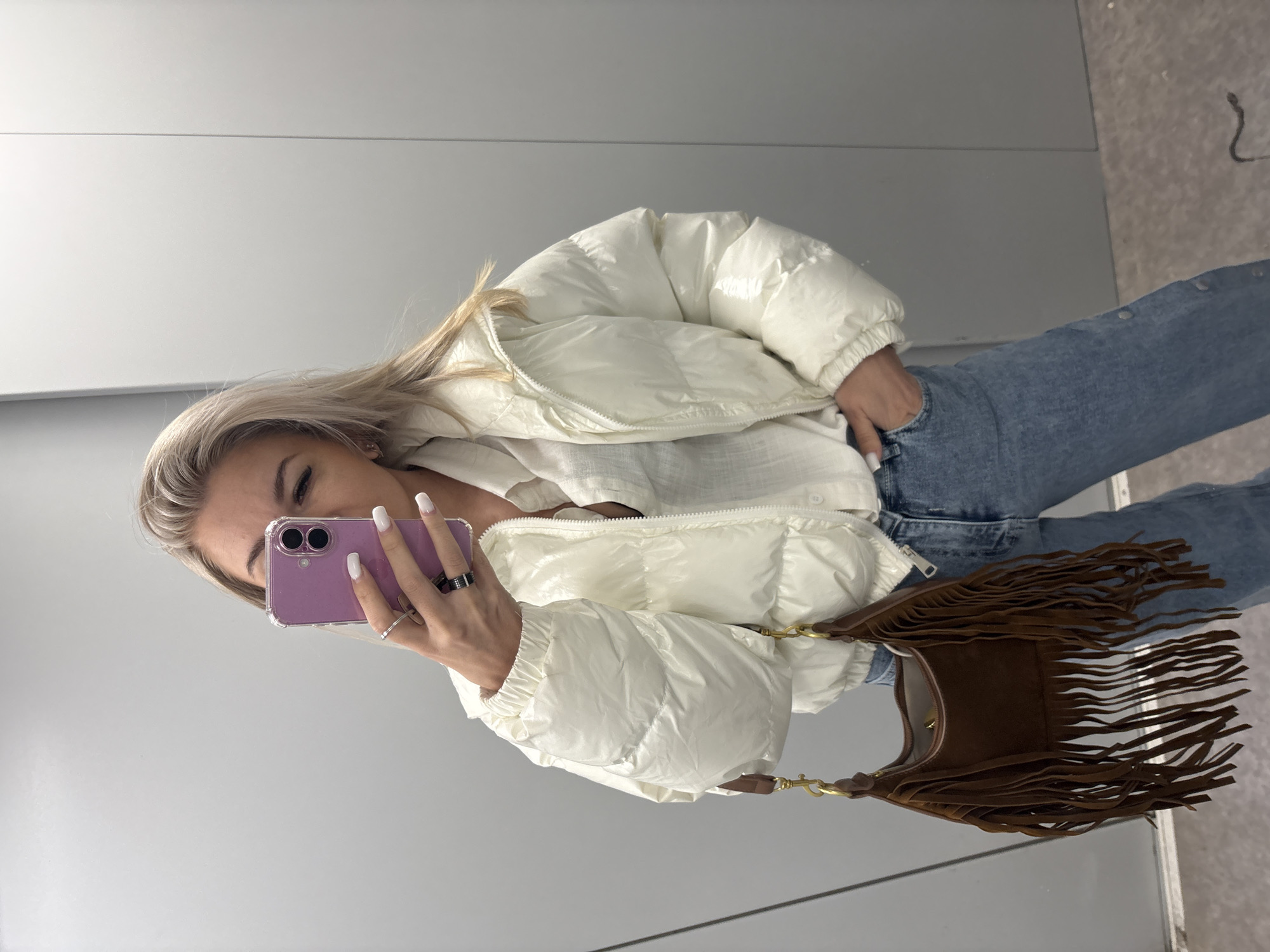 A young, fair-skinned woman with blonde hair is taking a selfie in what appears to be a dressing room or fitting room. She is wearing a white, puffy jacket over a light-colored top and blue jeans with buttons down the side. She is holding a purple smartphone with both hands, partially obscuring her face. The woman has rings on her fingers and white nail polish. A brown suede bag with fringe is hanging from her shoulder. The background is a plain, light-colored wall with a horizontal seam. The image is rotated slightly. There is no indication of this image being taken in Krasnodar, Russia.