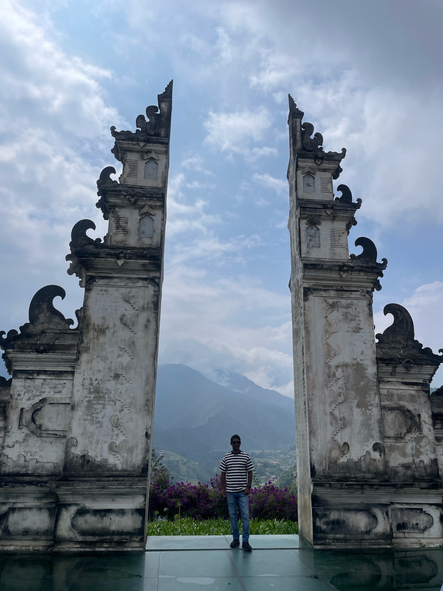 In Sa Pa Ward, Vietnam, a man stands centered within a grand, ornate stone archway. The archway is composed of two towering, weathered stone pillars, intricately carved with decorative motifs and topped with tiered, scalloped structures. The pillars are made of pale, aged stone, showing signs of wear and discoloration. Between the pillars, a hazy mountain landscape unfolds, with a town or village nestled in the valley below and lush green terraced hillsides climbing the slopes. In the foreground, a reflective surface, possibly a pool or polished floor, mirrors the scene. A vibrant band of purple flowers and green foliage runs across the middle ground, behind the man and in front of the distant landscape. The man is wearing a black and white striped t-shirt, blue jeans, sunglasses, and dark shoes. He is looking directly at the camera. The sky above is filled with scattered clouds, suggesting a partly cloudy day.