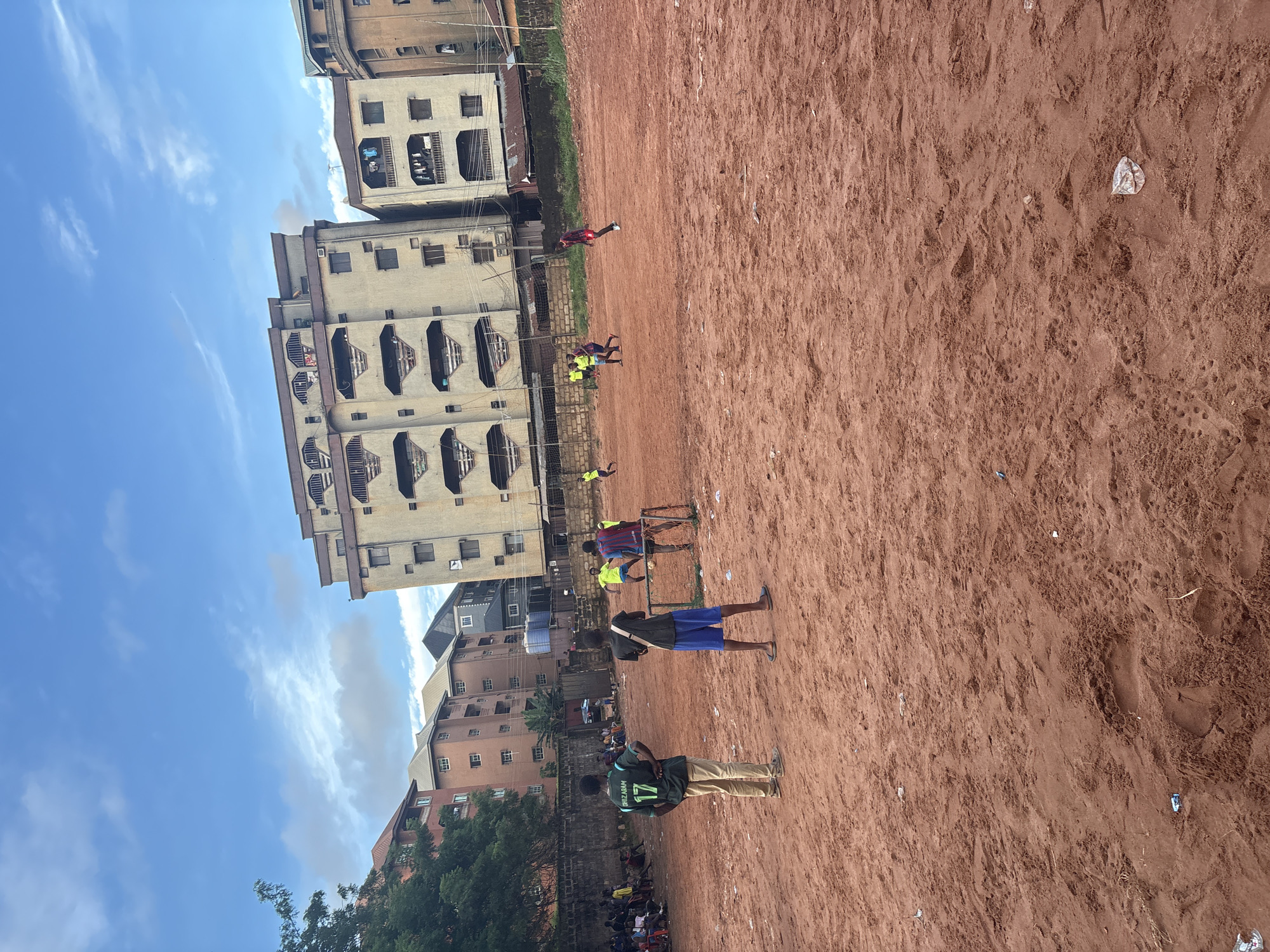 This is a photograph taken in Onitsha, Nigeria, depicting a group of young men playing soccer on a dirt field. The field is composed of reddish-brown soil, scattered with various pieces of litter. In the background, several multi-story buildings rise against a sky with scattered clouds. On the left side of the image, there is a brick wall and some trees. A rudimentary soccer goal is visible in the mid-ground. Several individuals are actively participating in the game, while others appear to be observing from the sidelines, some seated on chairs. The overall scene captures a moment of informal sports activity within an urban environment.