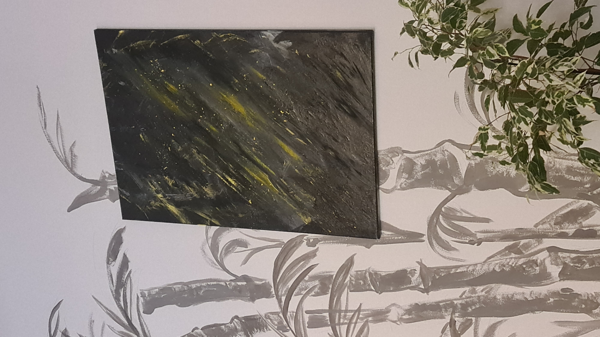 A black and yellow abstract painting hangs on a white wall with gray bamboo-like designs, next to a plant with green and white leaves. The location is not discernible from the image.