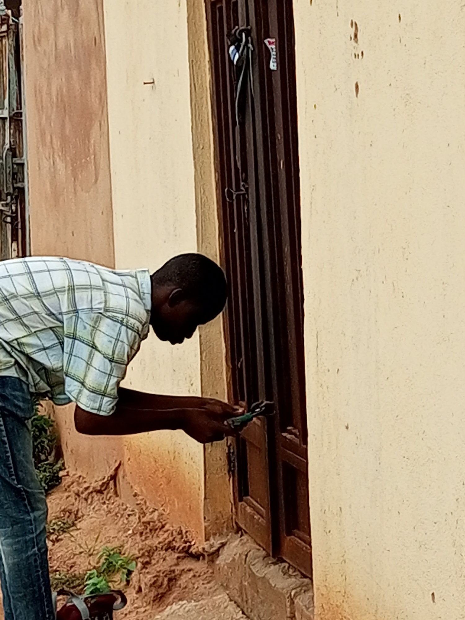 A young Black man wearing a plaid shirt and jeans is attempting to open a brown metal door with a pair of green pliers. He is standing in front of a building with a beige-colored wall on the right and a salmon-colored wall on the left. The ground is covered with dirt and small plants. The scene appears to be in Tempure, Nigeria.
