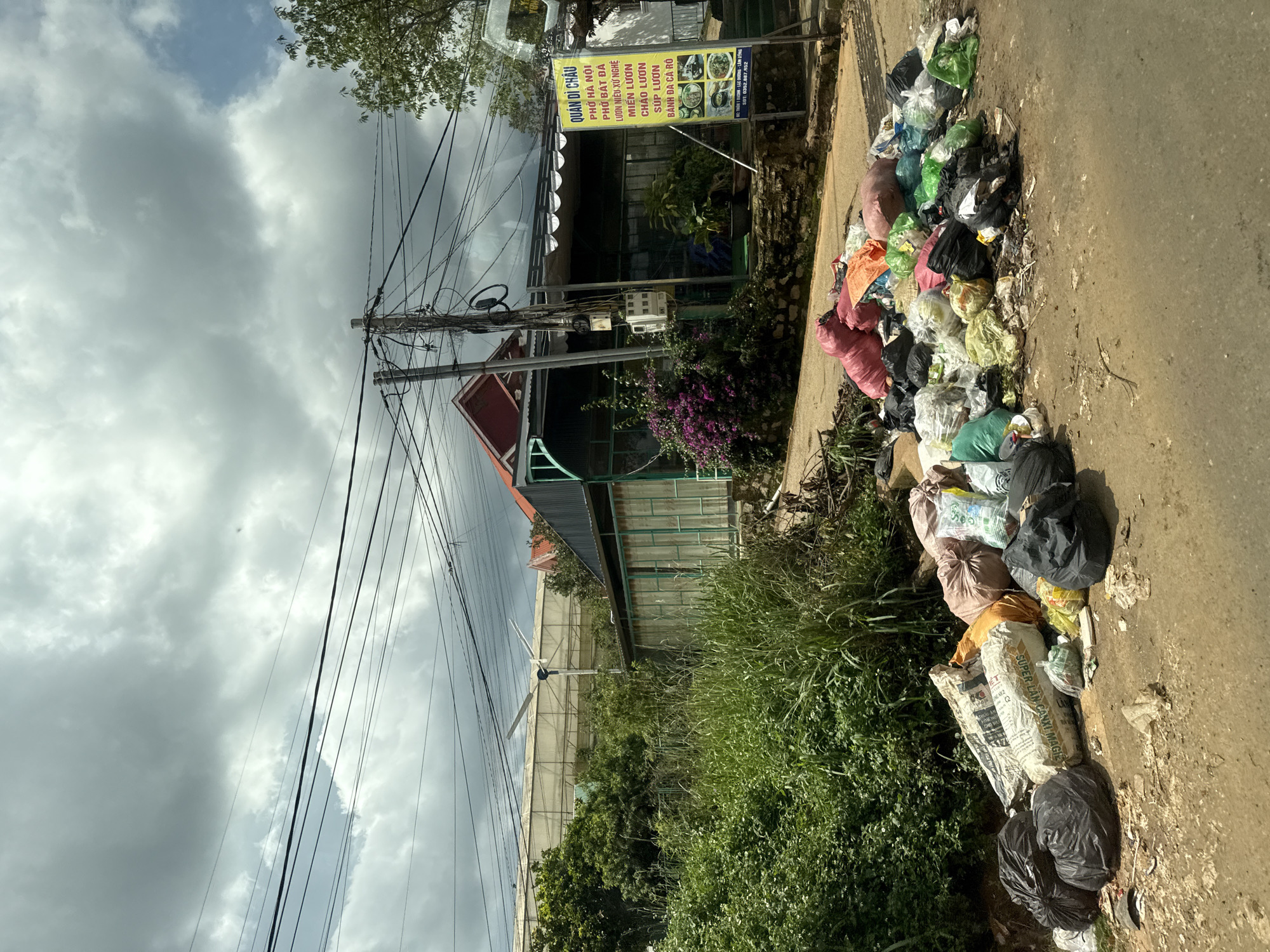 A pile of trash bags and loose garbage is accumulated on the side of a road in Thôn 1, Vietnam. The garbage includes black plastic bags, sacks of what appears to be cement or fertilizer, and various colorful plastic bags containing refuse. Behind the trash, there is a building with a green metal facade and a red roof, suggesting it might be a commercial establishment, possibly a restaurant, as a yellow sign with Vietnamese text is visible. Lush green vegetation and trees surround the building. Overhead, a complex network of electrical wires crisscrosses the sky, which is filled with heavy, gray clouds. In the distance, a greenhouse-like structure is visible, and a small wind turbine stands next to it. The road appears to be paved with asphalt and shows signs of wear. The scene suggests a rural or semi-urban area.