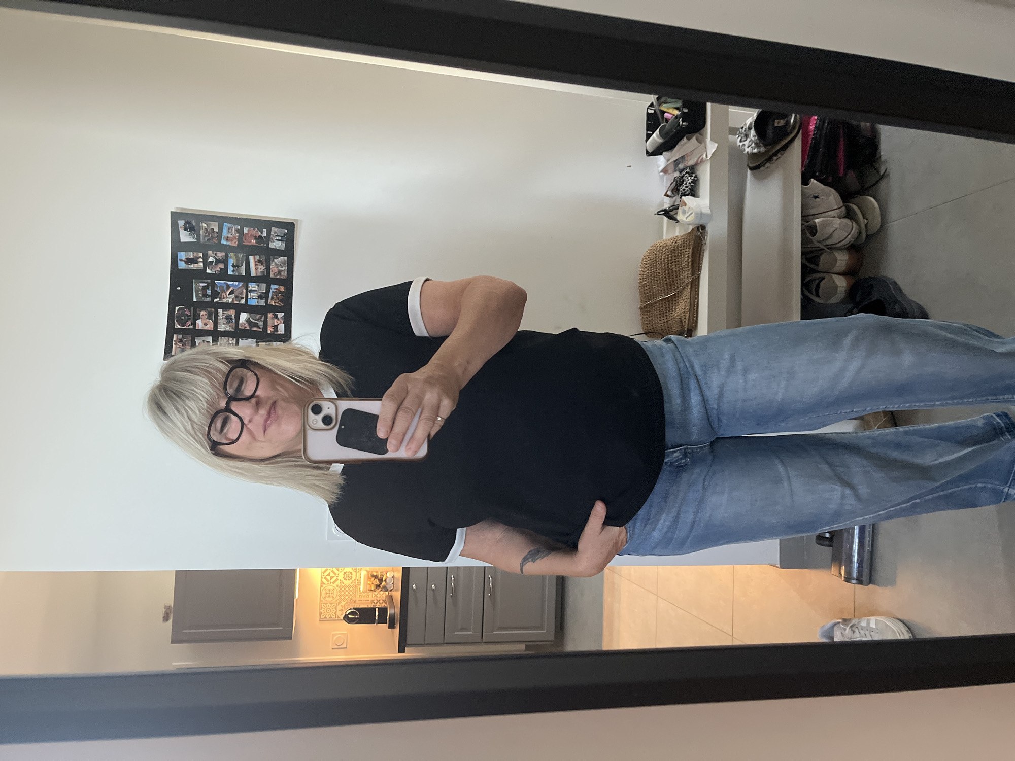 A woman is taking a mirror selfie in what appears to be a home in Chambœuf, France. She has blonde hair and is wearing glasses, a black t-shirt with white trim, and light-wash wide-leg jeans. Her left hand is in her jeans pocket. A phone is in her right hand, angled to capture the reflection. In the background, a kitchen area with cabinets and a backsplash is visible to the left. A shelf with various items, including a straw purse, glasses, and a black bin with toiletries, is to the right. The floor is tiled, and a vacuum cleaner is visible in the reflection on the floor. A framed collage of photos is on the wall behind her.