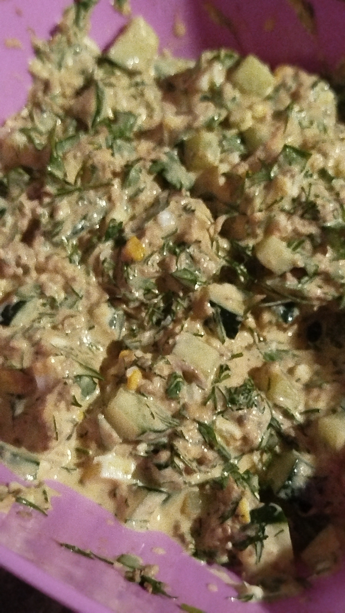 A close-up, top-down shot shows a close-up of a pink bowl filled with a chunky salad. The salad appears to be a mix of finely shredded meat, chopped cucumber or zucchini, possibly corn kernels, and fresh dill. A creamy dressing coats the ingredients, giving it a slightly wet and clumpy texture. The lighting is somewhat dim and appears to be artificial. The image does not provide any information that could be used to determine its location in Kostiantynivka, Ukraine, or any other specific location.
