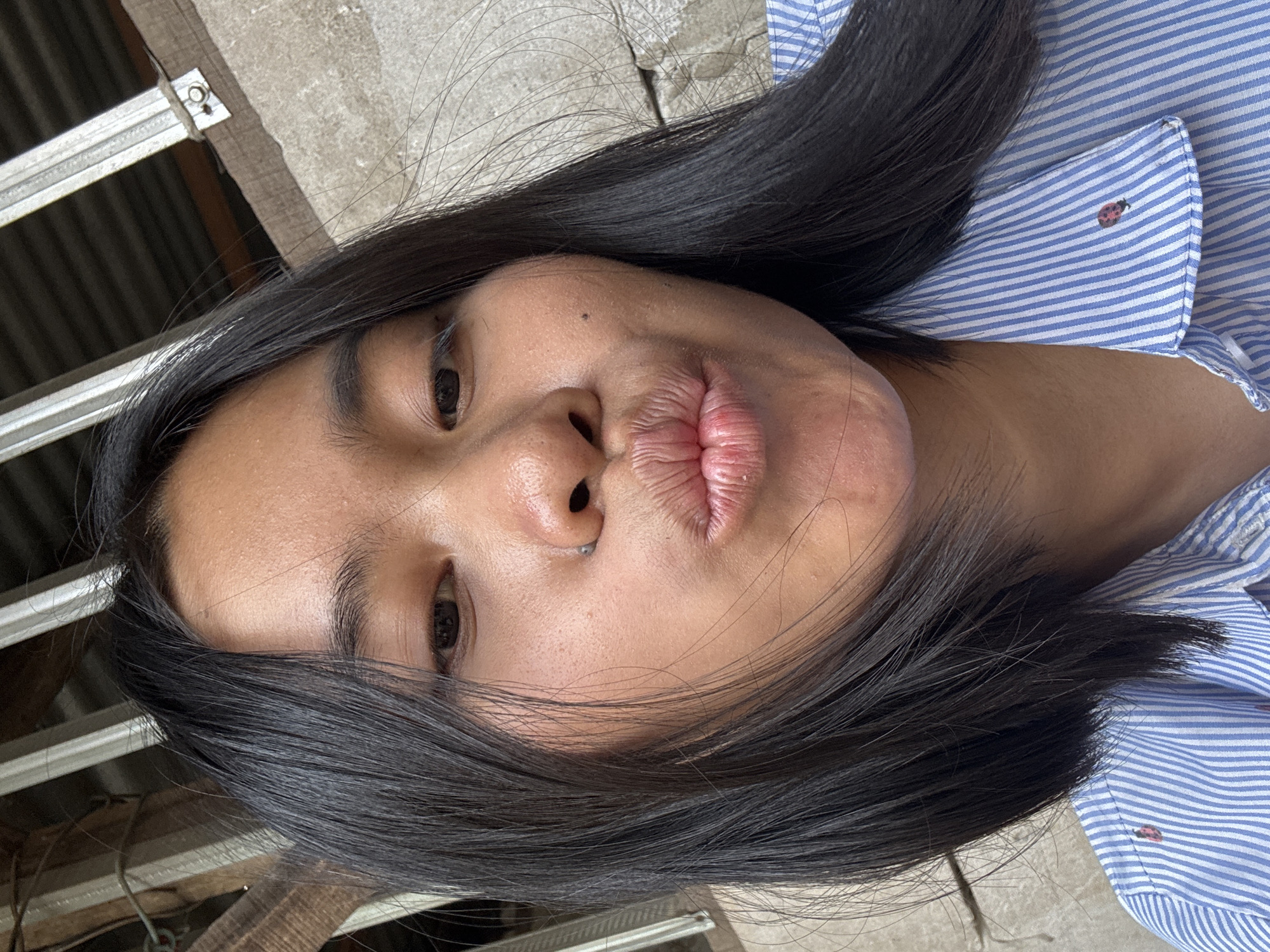 A close-up, eye-level, outdoor selfie shows a young woman with dark hair and light brown skin puckering her lips. She is wearing a blue and white striped button-down shirt. The background consists of a textured concrete wall and what appears to be part of a roof structure with corrugated metal and wooden beams. The image was taken in East Jakarta, Indonesia.