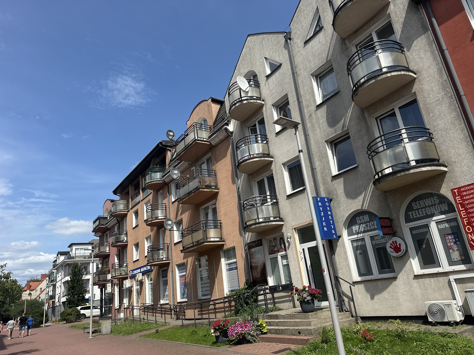 The image shows a street scene in Pruszcz Gdański, Poland, on a sunny day with a blue sky and scattered clouds. In the foreground, a red brick sidewalk lines the street. To the right of the sidewalk is a strip of grass with flower pots containing colorful flowers. Further to the right is a stepped entrance to a building.
The main subject of the image is a row of multi-story buildings lining the street. The buildings have a modern architectural style, with balconies on each floor. The building in the foreground on the right is light gray and has several windows and balconies. A white air conditioning unit is visible near the bottom of this building. Next to it is a building with a terracotta-colored facade, also featuring balconies and windows. To the left, the buildings continue with different colors and designs, including white and darker tones.
Several signs are visible on the buildings, indicating businesses. One sign on the terracotta-colored building reads "CENTRUM MEDYCZNE" (Medical Center). On the gray building, signs include "SERWIS TELEFONÓW" (Phone Service), "GSM PRUSZCZ" (GSM Pruszcz), and "FRYZJER KOSMETYKA" (Hairdresser Cosmetics).
In the background, more buildings and trees are visible, suggesting a residential or commercial area. A few people are walking on the sidewalk in the distance, moving away from the camera. A white car is parked on the street. Satellite dishes are mounted on some of the balconies. The overall impression is a well-maintained urban environment.