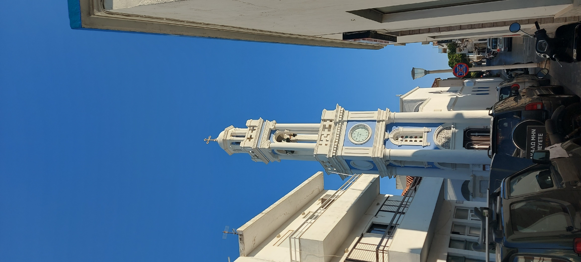 The image shows a narrow street in the Municipal Unit of Rhodes, Greece. A white and blue bell tower with a clock is the most prominent structure, topped with a cross. Adjacent to the bell tower is a white building with balconies. On the right side of the street, a white building partially obscures the view. Cars and a scooter are parked along the street. The sky is clear blue. A street lamp and a signpost are also visible.