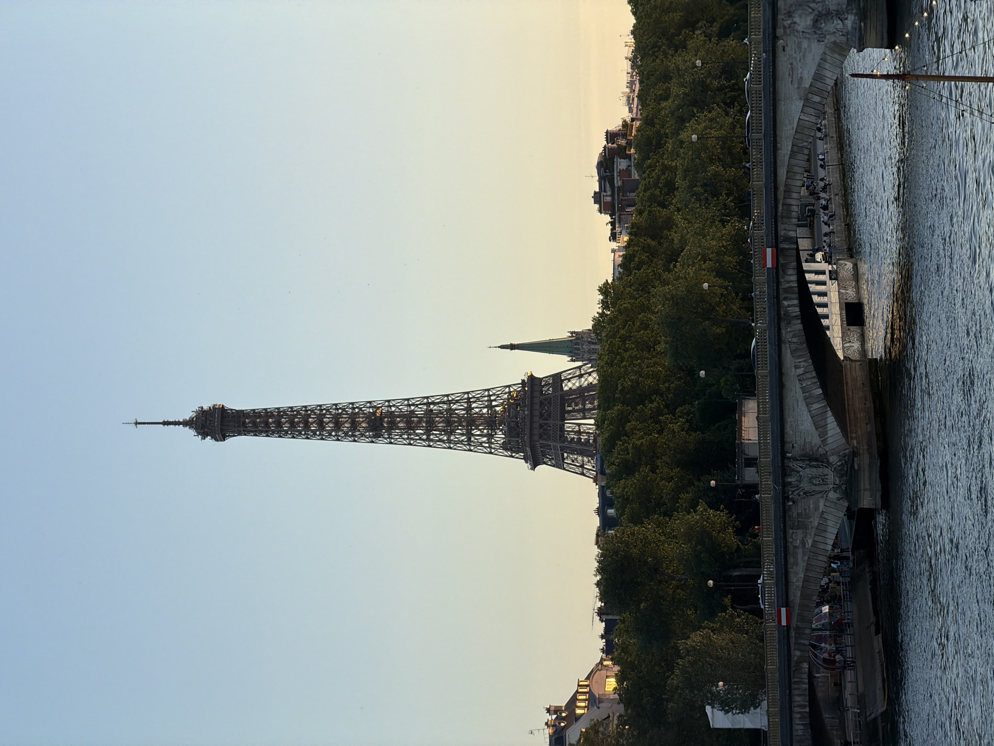 The image shows the Eiffel Tower in Paris, France, rising prominently against a pale sky. The tower is framed by trees and buildings lining the Seine River. A bridge extends over the river, with people visible on its steps, suggesting a gathering or leisurely activity. The water surface is textured with ripples, reflecting the ambient light.