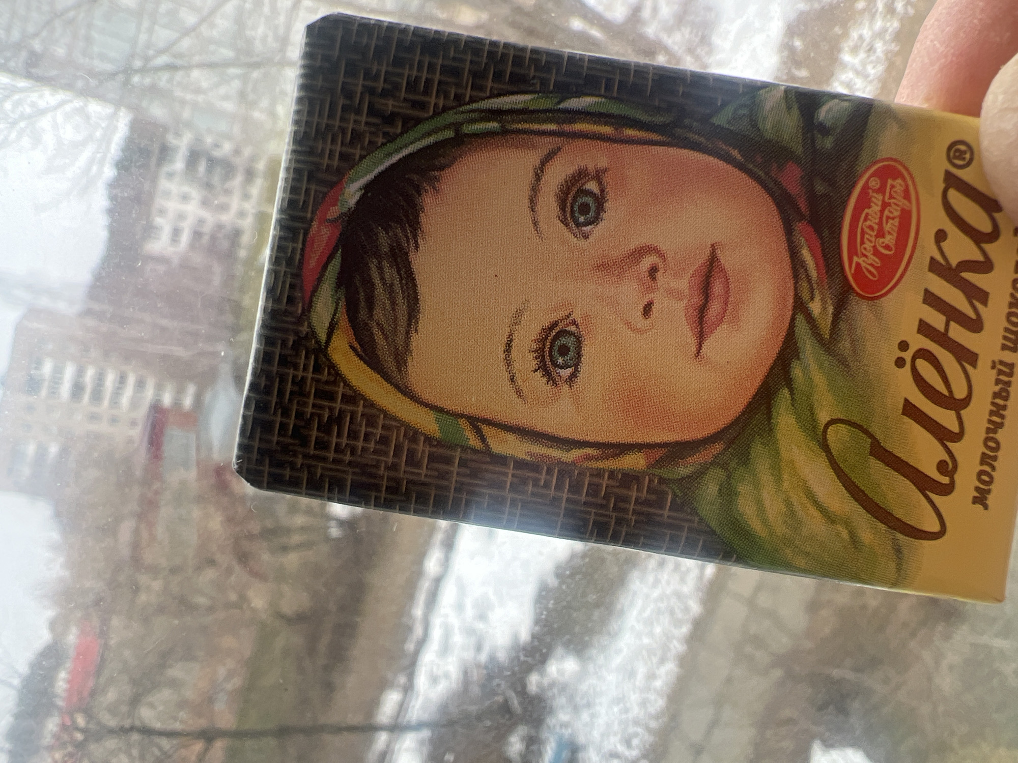 Here is a description of the image:
A partially rotated close-up shot shows a rectangular chocolate bar wrapper with a portrait of a young girl. The girl has light skin, a small nose, and light eyes. She is wearing a headscarf. The brand name "Alenka" in Cyrillic script is written in cursive on the right side of the wrapper. The background of the wrapper features a woven pattern. The wrapper is placed on a surface, possibly a windowsill, which is situated in front of a window. The blurred outdoor view shows buildings, trees, and a snowy landscape. This image was taken in Chaikovsky, Russia.