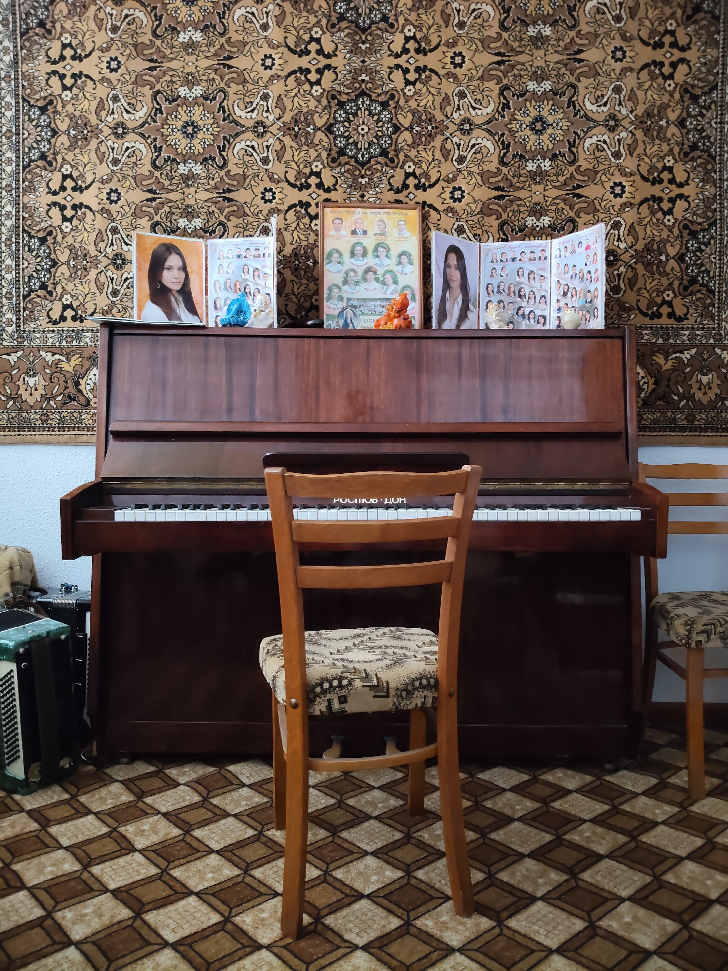 A medium shot captures a domestic scene featuring an upright wooden piano with an open lid, displaying photographs and decorative items on its surface. A wooden chair with a patterned seat sits directly in front of the piano. To the left of the piano, an accordion is visible. The background is dominated by a large, ornate rug with intricate brown and beige patterns, hanging on the wall. The floor is covered with a patterned carpet in shades of brown and beige, creating a checkered effect. The scene appears to be indoors, likely a living room or music room. The photograph provides a clear view of the piano, chair, accordion, and the decorative elements, offering a glimpse into the room's ambiance.