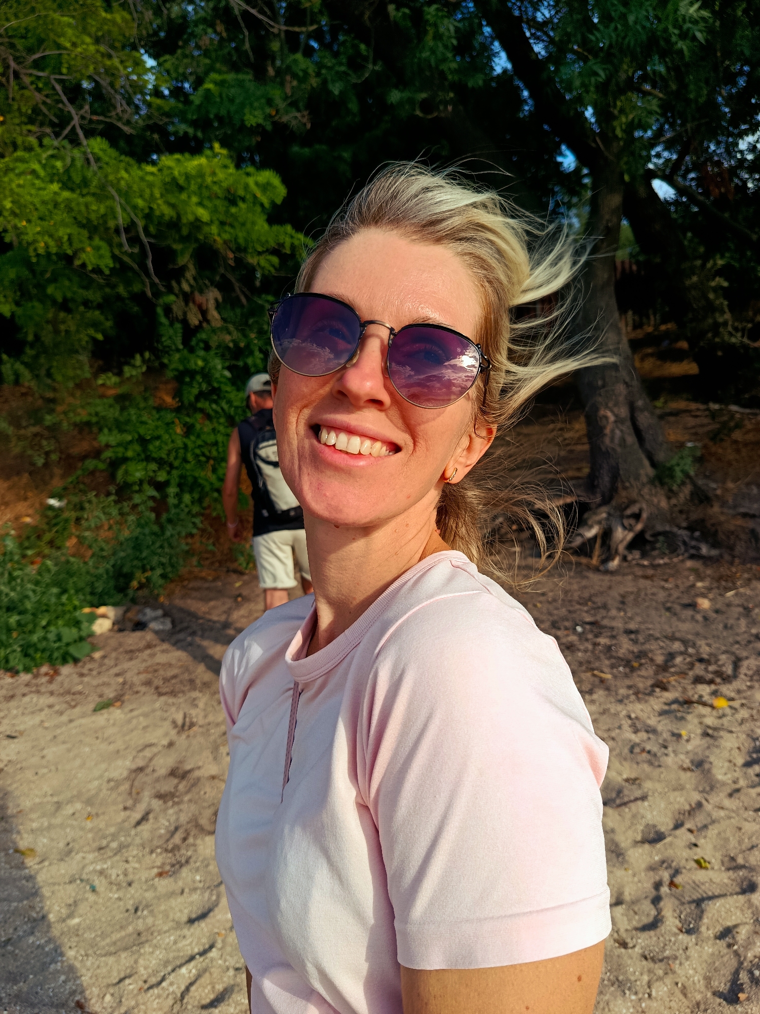 A woman with blonde hair and sunglasses is smiling at the camera. She is wearing a light pink t-shirt. In the background, a man is walking away from the camera. The scene appears to be a sandy beach with trees and foliage. The lighting suggests it is daytime, possibly late afternoon, due to the warm tones and shadows. The location is described as Unknown, Unknown.